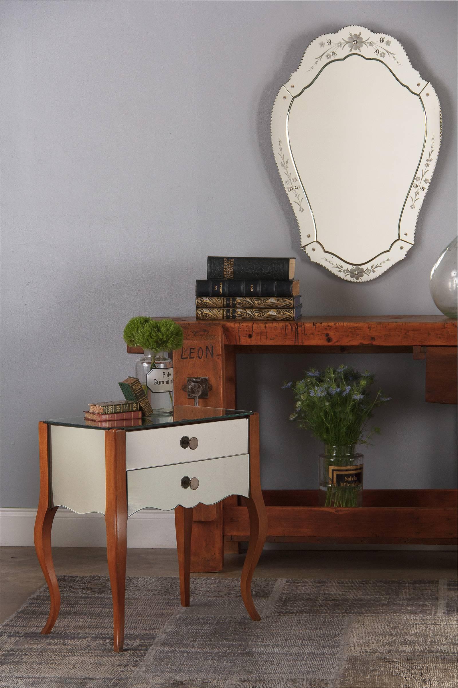 A petite midcentury Venetian glass mirrored chest in blonde mahogany, 1950s. Two drawers with silver metal round pulls. Both sides and the front boast gracefully scalloped lower profiles. The rectangular mirrored top is enhanced by marquise and dot