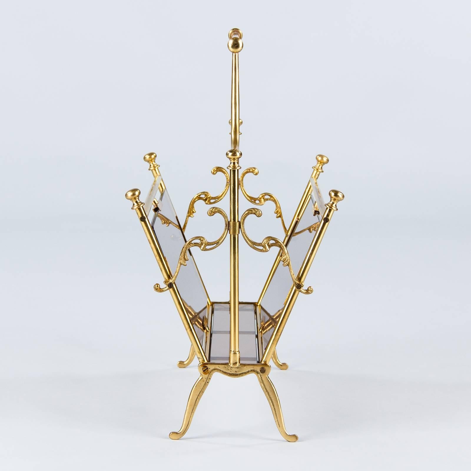 A 1950s magazine holder made of polished brass and smoked glass. Brass rods with round finial tops form the frame, which rest on slightly curved, tapered legs with incised decoration. Flourish shaped brass pieces with pierced detailing join the