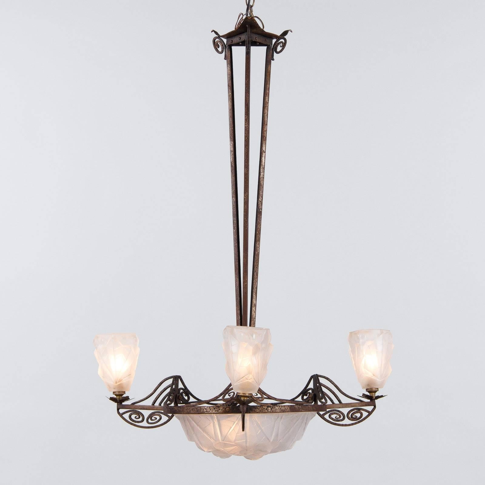 A striking French Art Deco chandelier in iron and frosted cut glass, signed by Degue, circa 1930. The centerpiece is a large bowl of frosted glass cut into numerous angles, with three stylized rosettes and their leaves being framed by more subtle