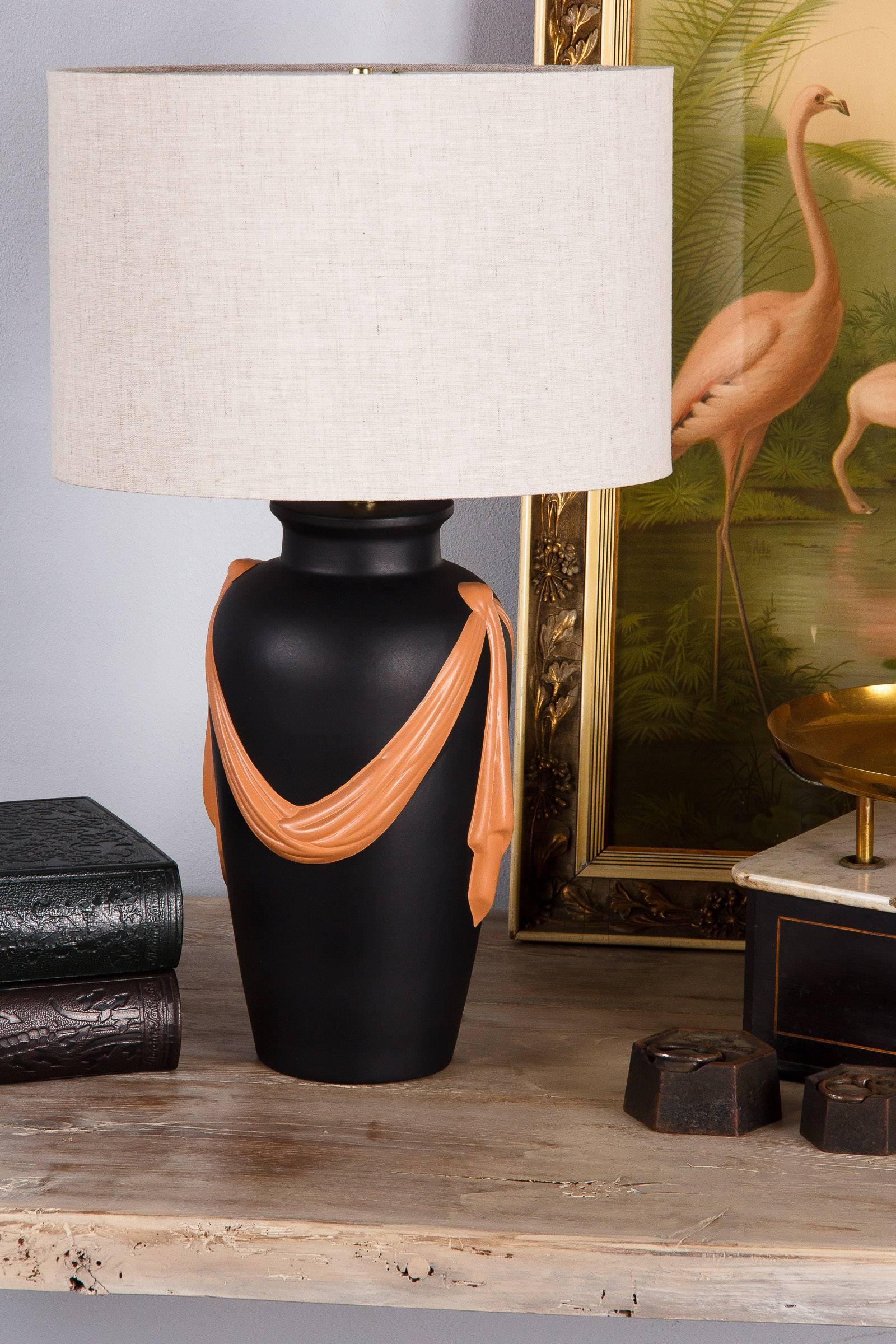 A black terracotta lamp decorated with a sash in salmon tones. The lamp is housing a three way light bulb. Tailor made lampshade is 16
