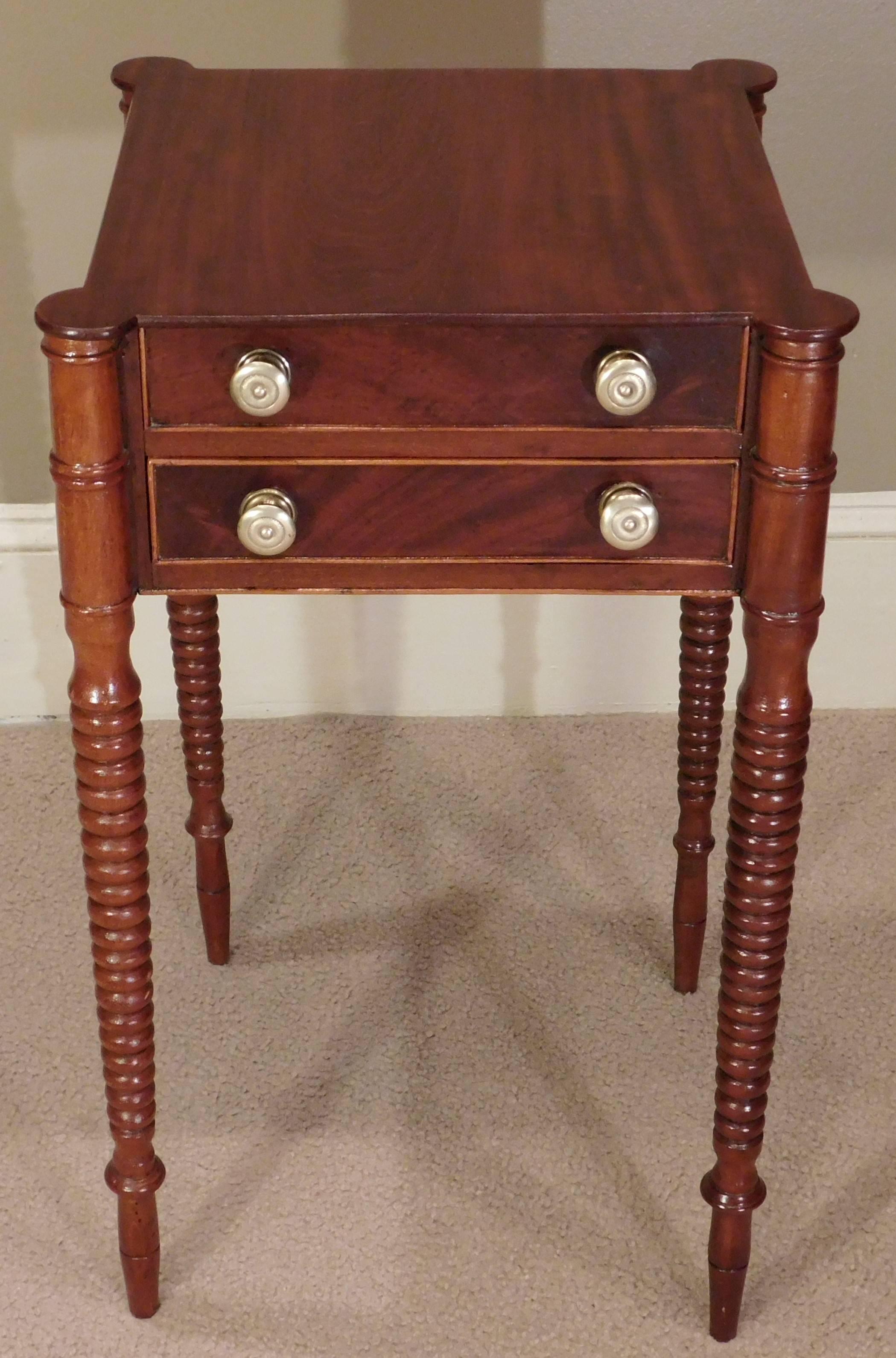 This Sheraton work table is figured mahogany and cherry with poplar secondary wood and old replaced solid brass pulls. The stand is finished on all four sides with turned turret corners above spiral turned legs.