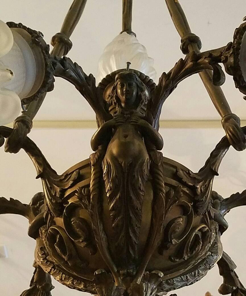 Bronze six branch chandelier with six ruffled satin glass shades and centering a flame-form shade on the torch, the candle branches in the arms of a caryatid figure.