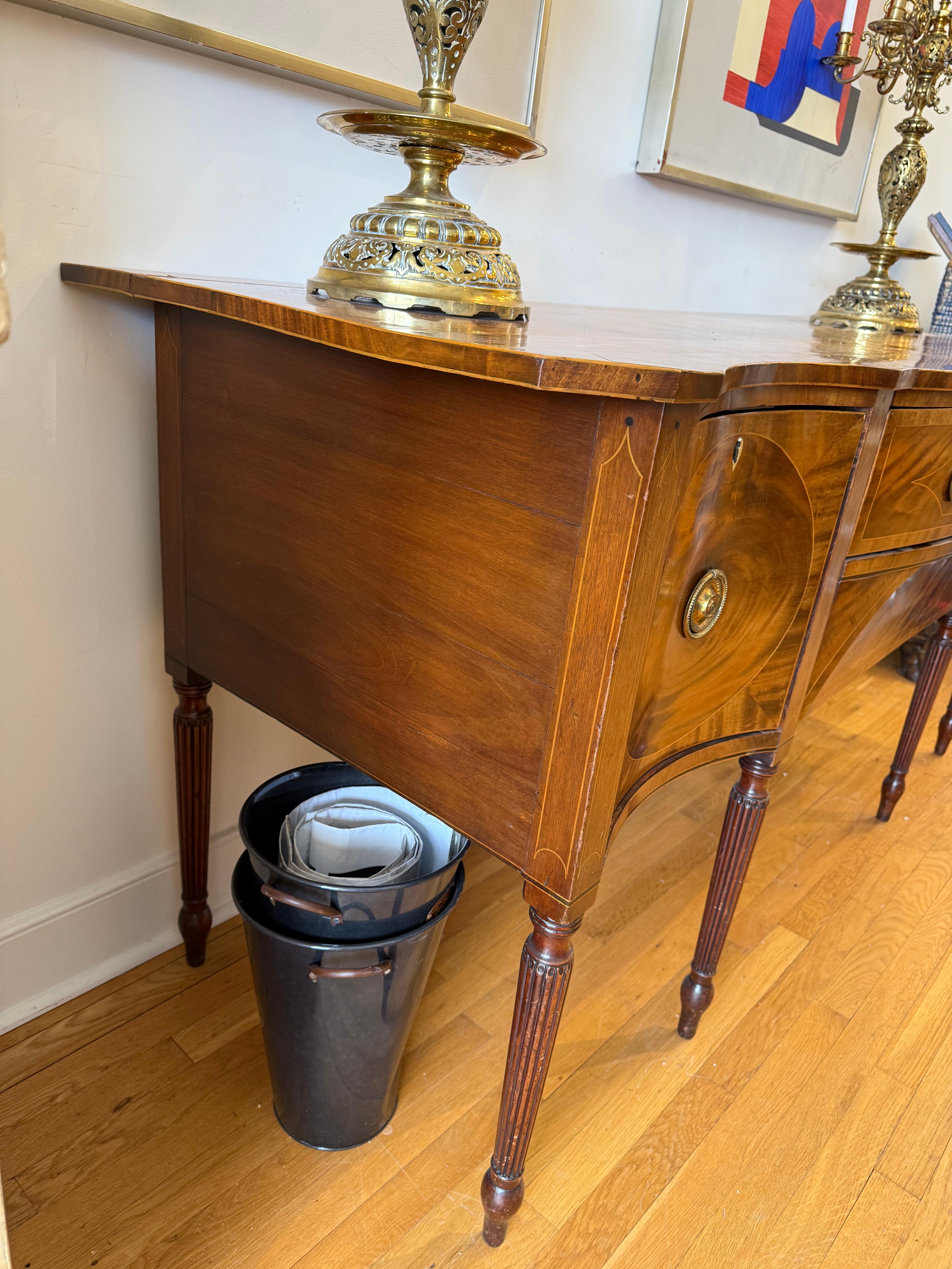 A masterpiece of Georgian elegance, this early Sheraton period serpentine sideboard exemplifies the refined craftsmanship and architectural harmony of late 18th-century England. Made circa 1790, this commanding piece is fashioned from radiant flame