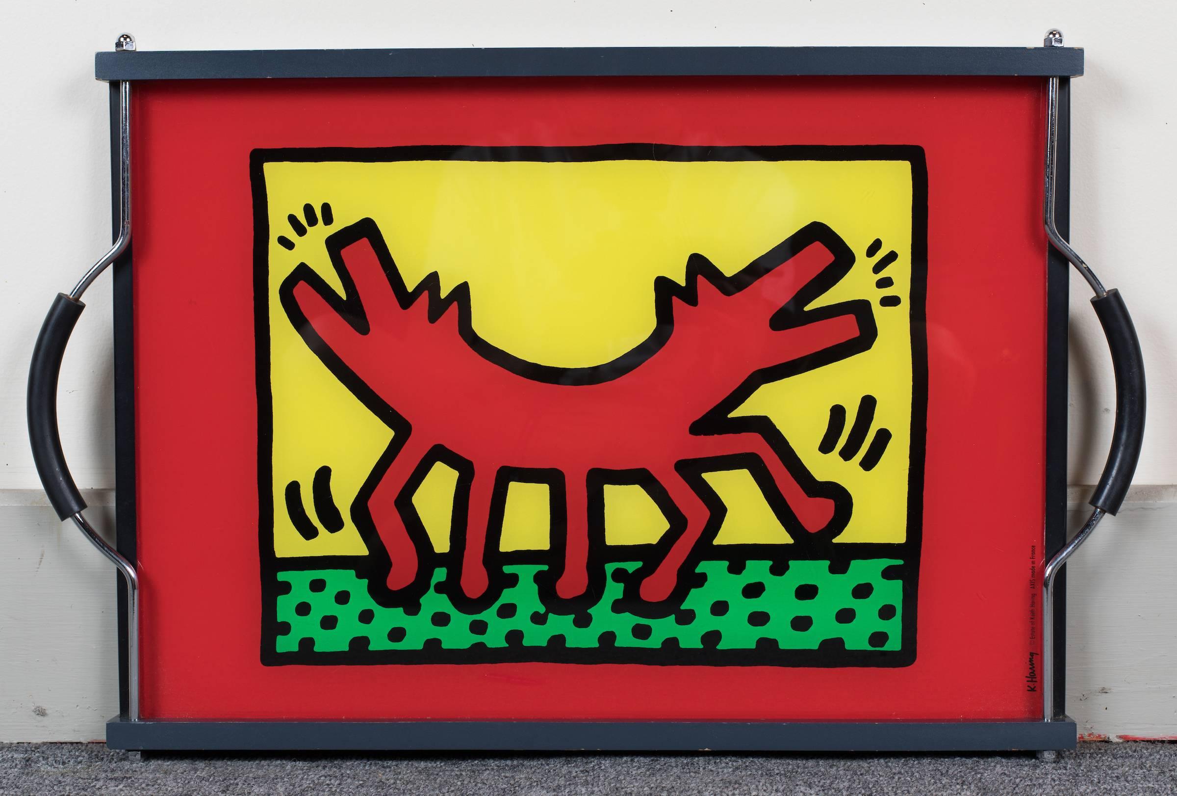 Whimsical Keith Haring eglomisé glass serving tray showing a double headed barking dog, fitted with chrome and rubber handles, the glass held with painted wood supports.
Signed: K. Haring, estate of Keith Haring, AXIS made in France.