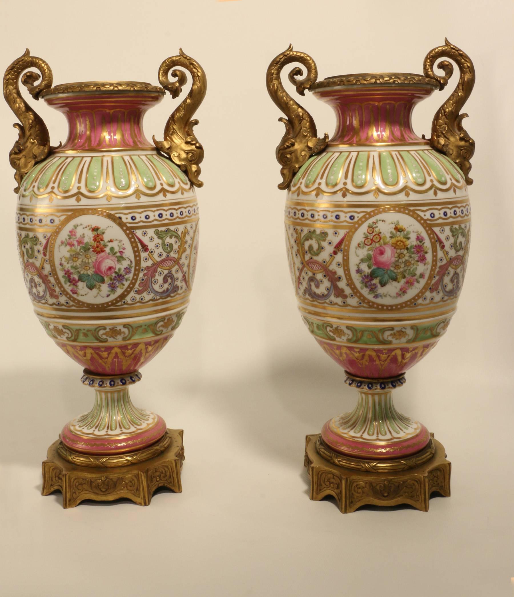 A pair of French Sevres style porcelain urns, each of baluster form, moulded and painted in pompadour pink and eau de nil and gilt, on a white ground. Each is painted with alternating jeweled oval panels of floral and musical trophies amid stylised