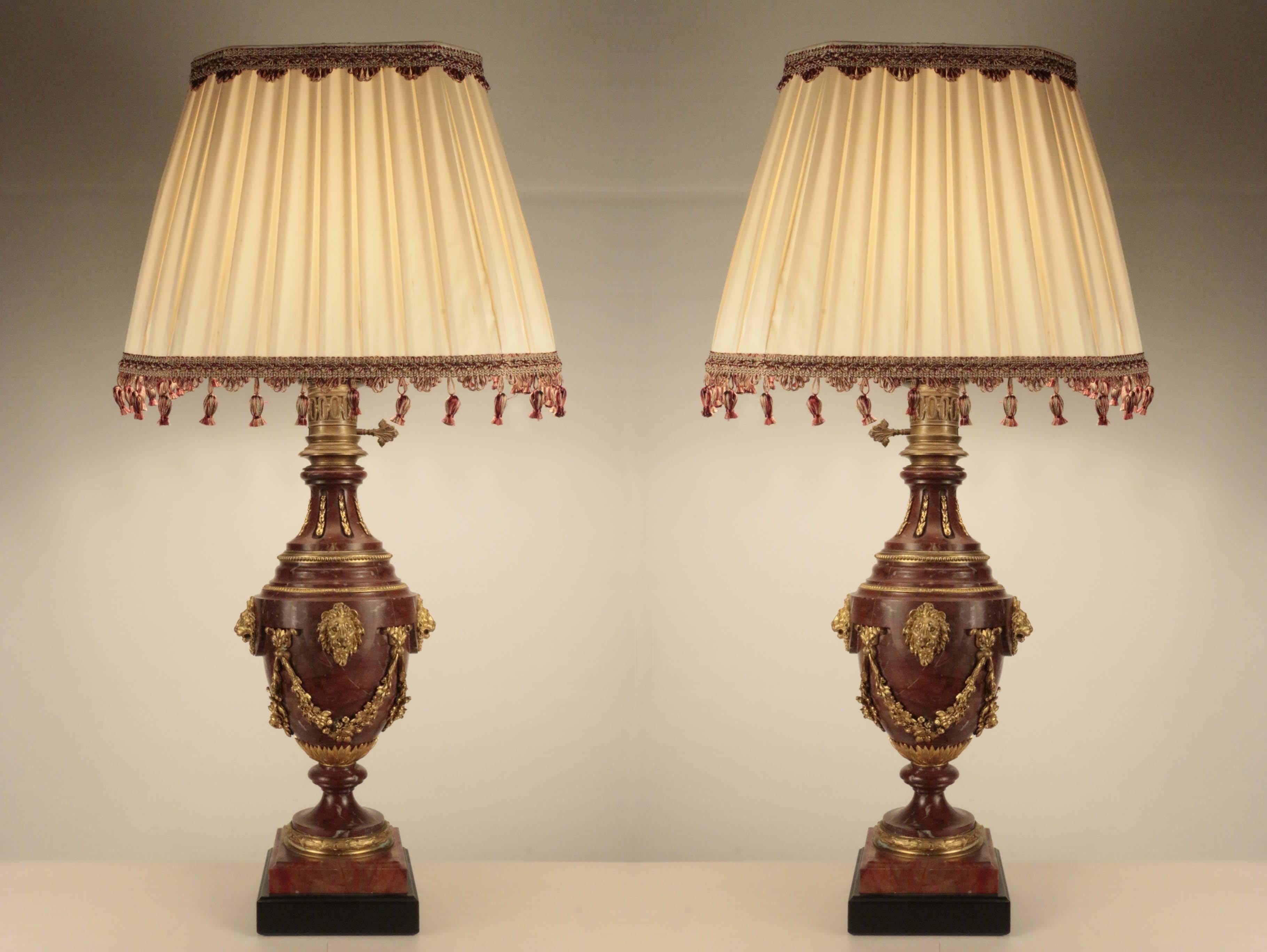 Pair of rouge marble and gilt bronze baluster shaped oil lamps, later fitted for electricity, the necks and bodies decorated with gilt bronze banding and garlands and accented with lions' heads; supported on marble socles on ebonized wood bases.