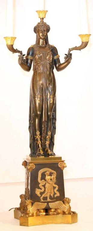 
Crisply modelled as a standing godess, supporting ebonised and gilt arms, her slender body clad in a clinging toga with gilt tassels , arm bands, and sandals. She stands on an elaborate tapering square base base applied with gilt mounts and raised