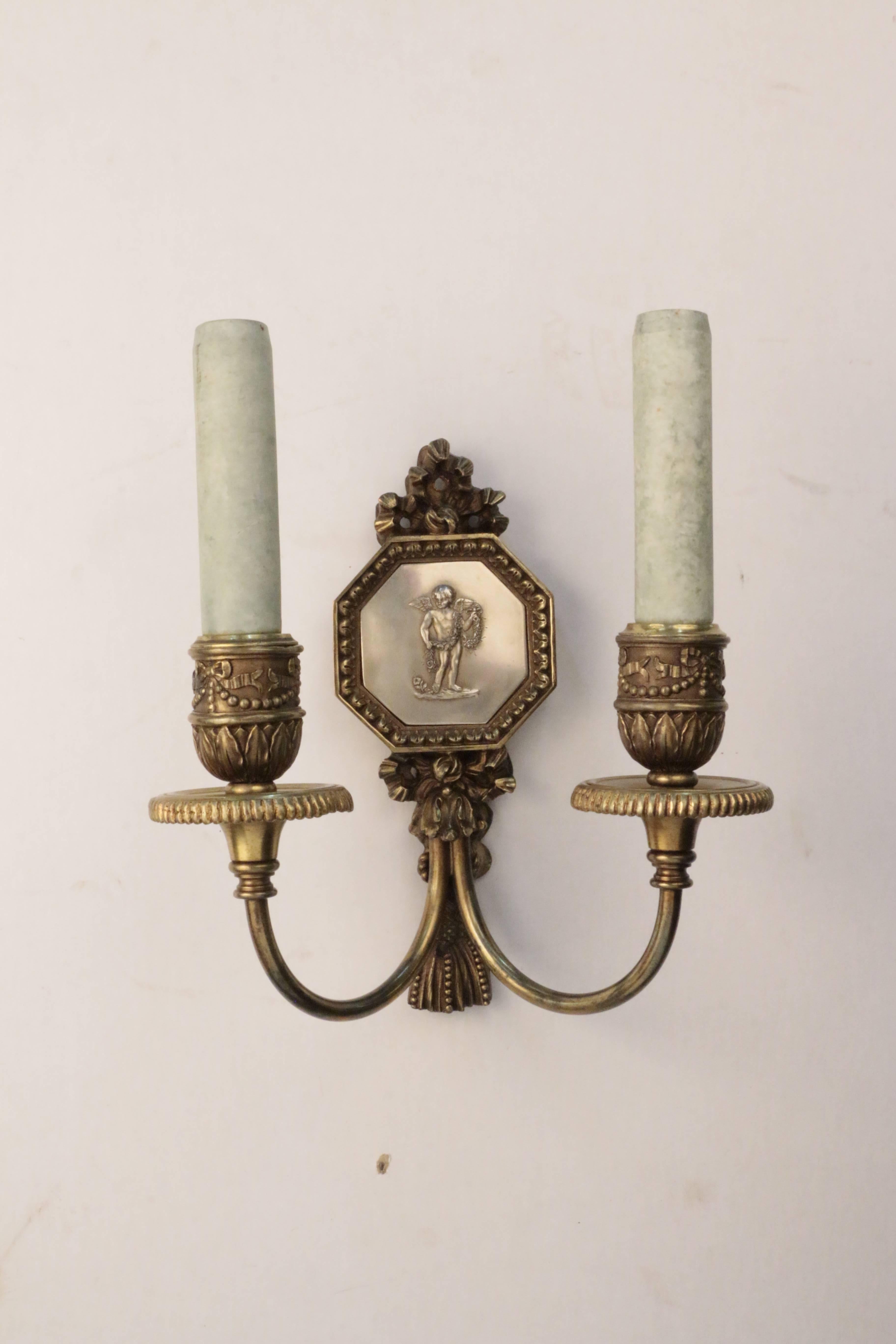 Pair of E F Caldwell bronze two-light wall sconces, each crisply modeled in Louis XV1 style with acanthus, beaded and ribbon motif and set with an hexagonal silvered panel depicting a cherub with floral garland. The bisque candle sleeves are