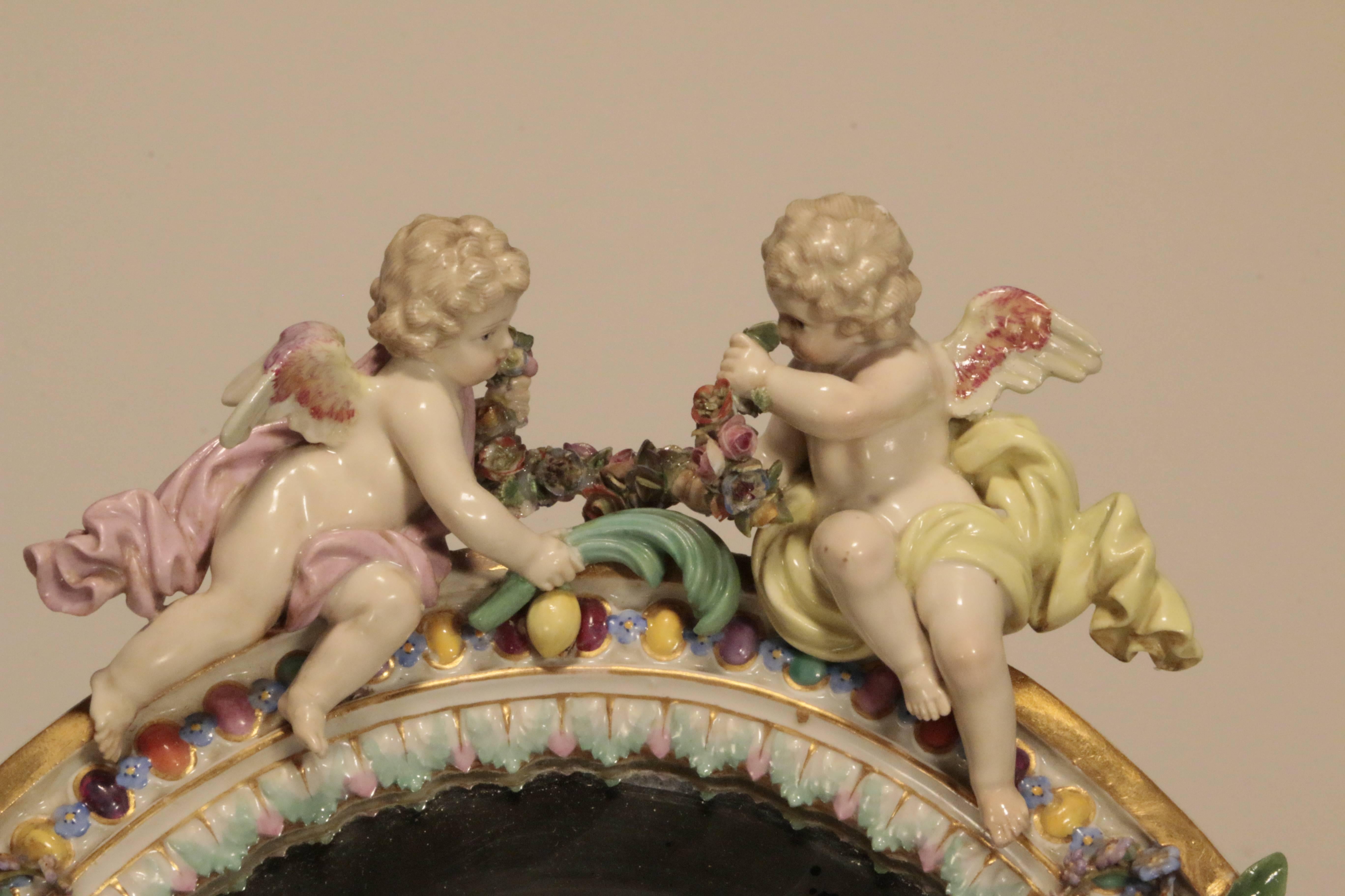 An antique oval Meissen table mirror crested with two cherubs and a garland, with beveled mirror, the frame hand-painted with fruit and applied leaves and flowers, with strut panel, fully marked circa 1870.
