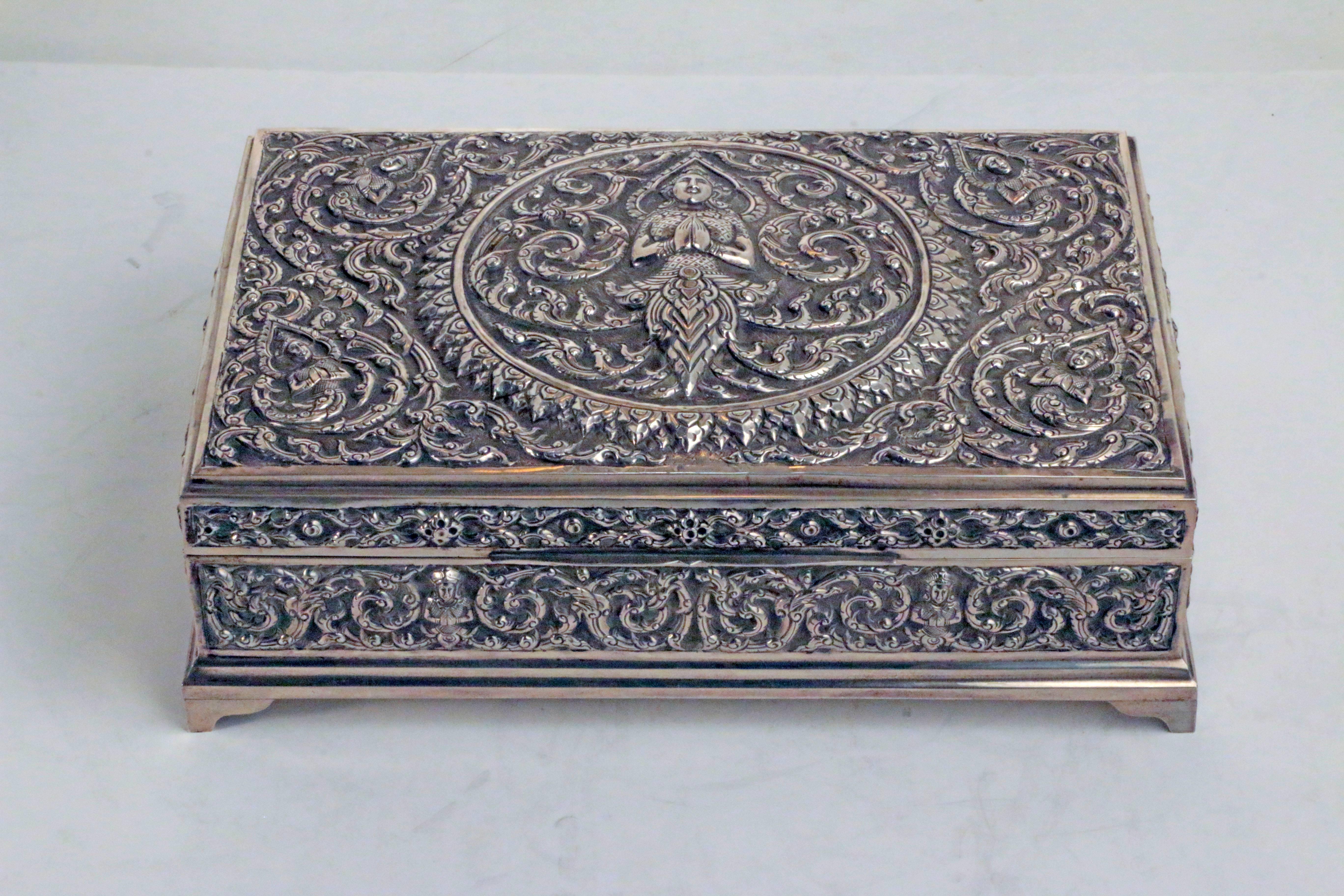 A sumptuous sterling silver cigar box, from old Siam, centred by a raised representation of the Buddha his hands in a mudra of calm compassion and welcome. The spandrils of the box each contain a single figure with hands similarly clasped amid the