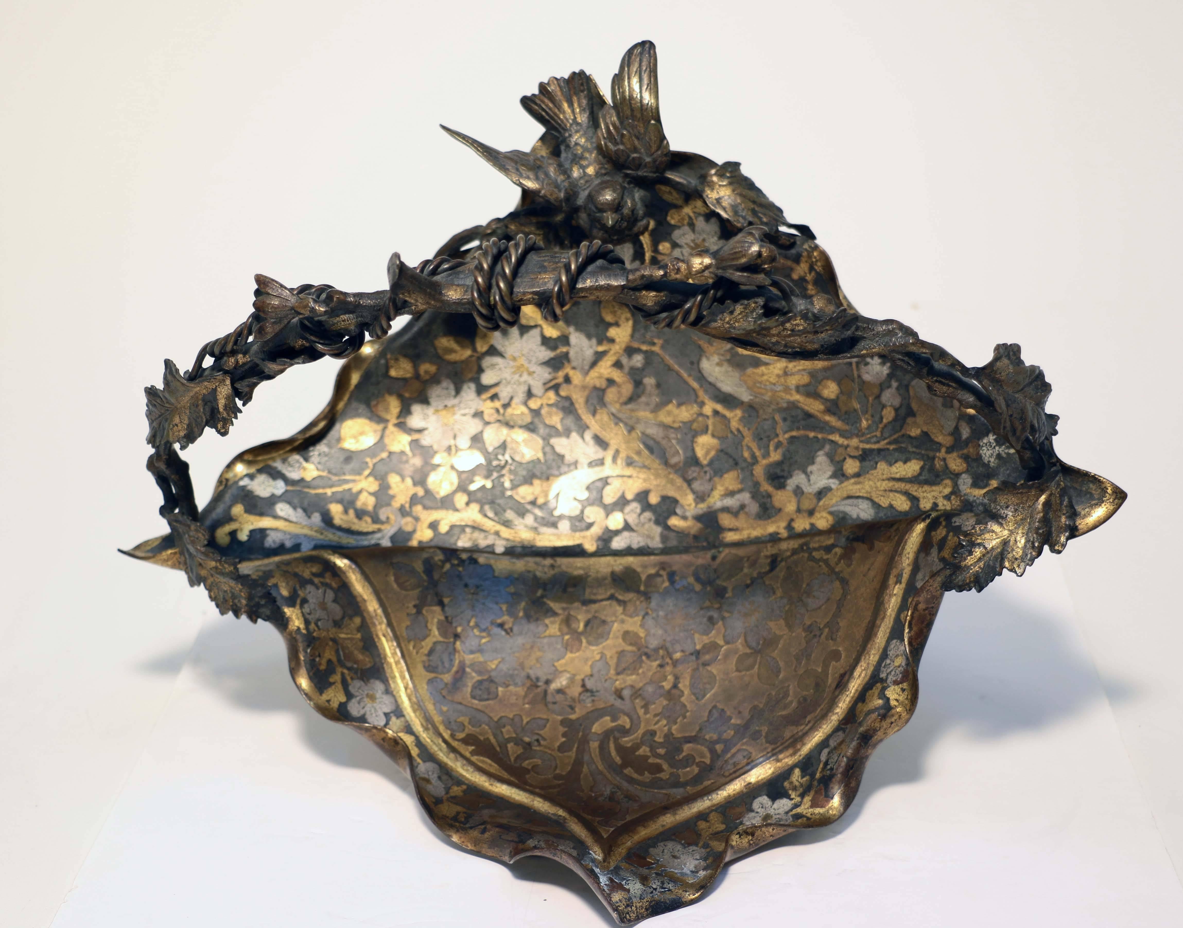 An American Aesthetic Movement Basket, naturalistically modeled as a bird perched on a a leafy twig, above a shallow dish simulating cloth, decorated with gilt and silvered leaves and flower heads, the handle modeled as a bent twig with simulated