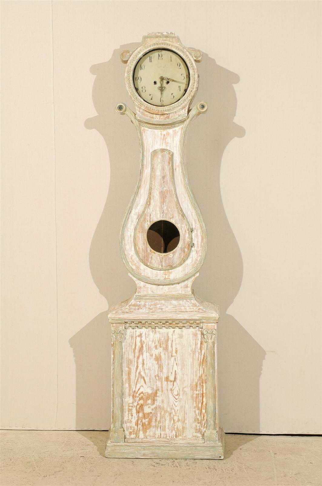 Eine schwedische Uhr aus bemaltem Holz aus dem 19. Jahrhundert mit Voluten auf beiden Seiten des Kopfes. Diese Uhr hat einen leierförmigen Korpus und einen rechteckigen Sockel, der von zwei stilisierten korinthischen Säulen flankiert wird. Diese Uhr