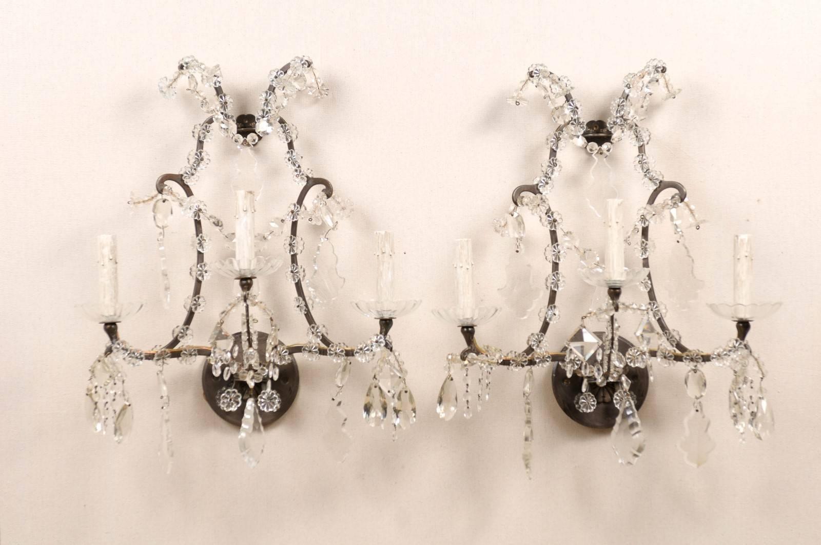 A pair of crystal three-light sconces. This pair of Italian vintage sconces is made of a scrolled armature decorated with small size crystal flowers and glass bobèches. The sconces are also decorated with various faceted crystals. The back plate