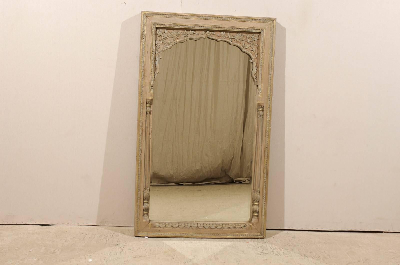 A vintage British Colonial style mirror. This mirror features a natural color carved wood surround with light grey and gold painted details throughout. The carvings feature pretty floral motifs. The sides of the mirror are flanked with carved thin