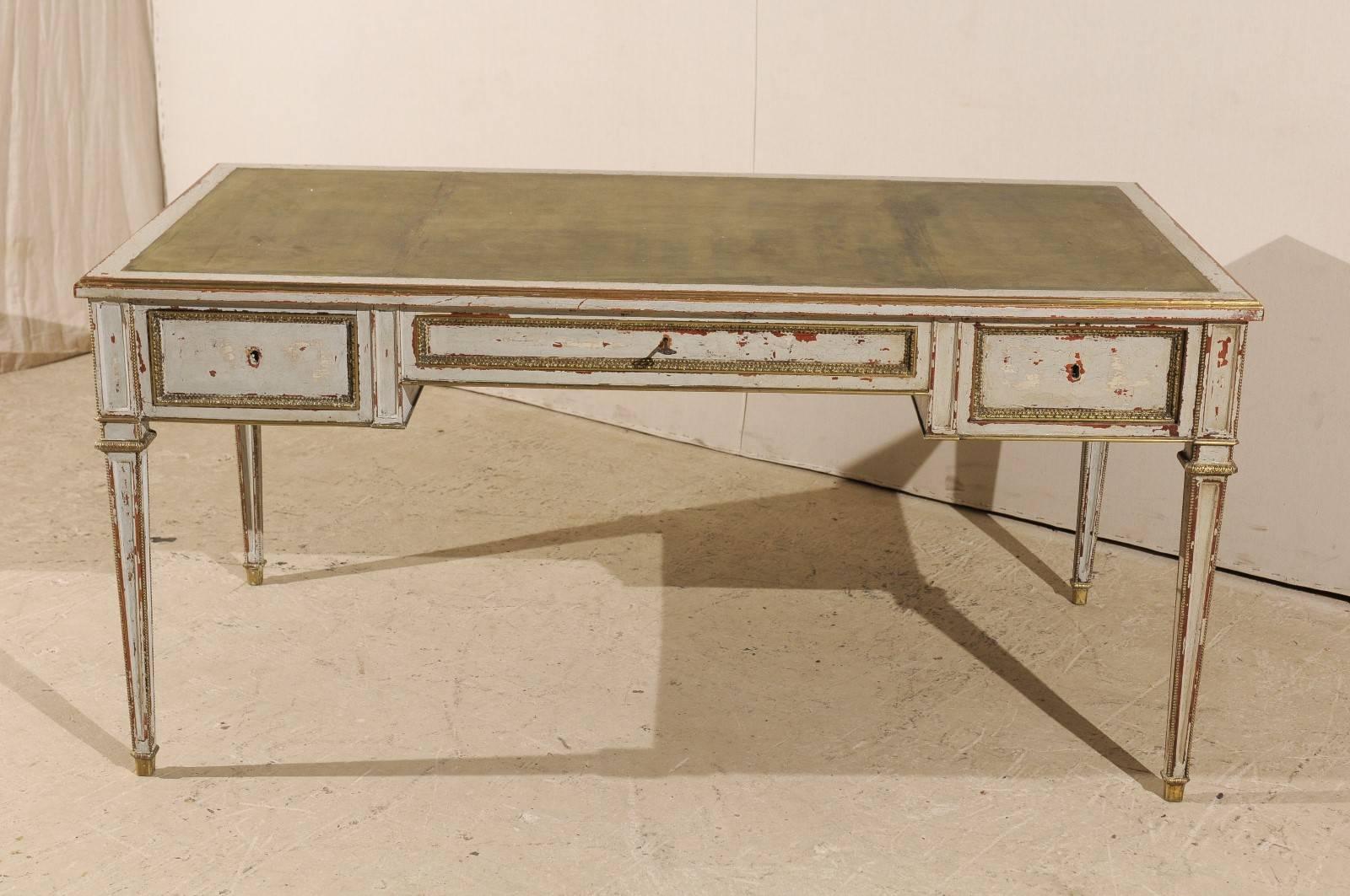 A French painted wood desk. This French three-drawer desk from the 20th century features a green leather top that has embossed edges. The desk is raised on four tapered legs. This piece is grey-white throughout with some crackled paint showing a red