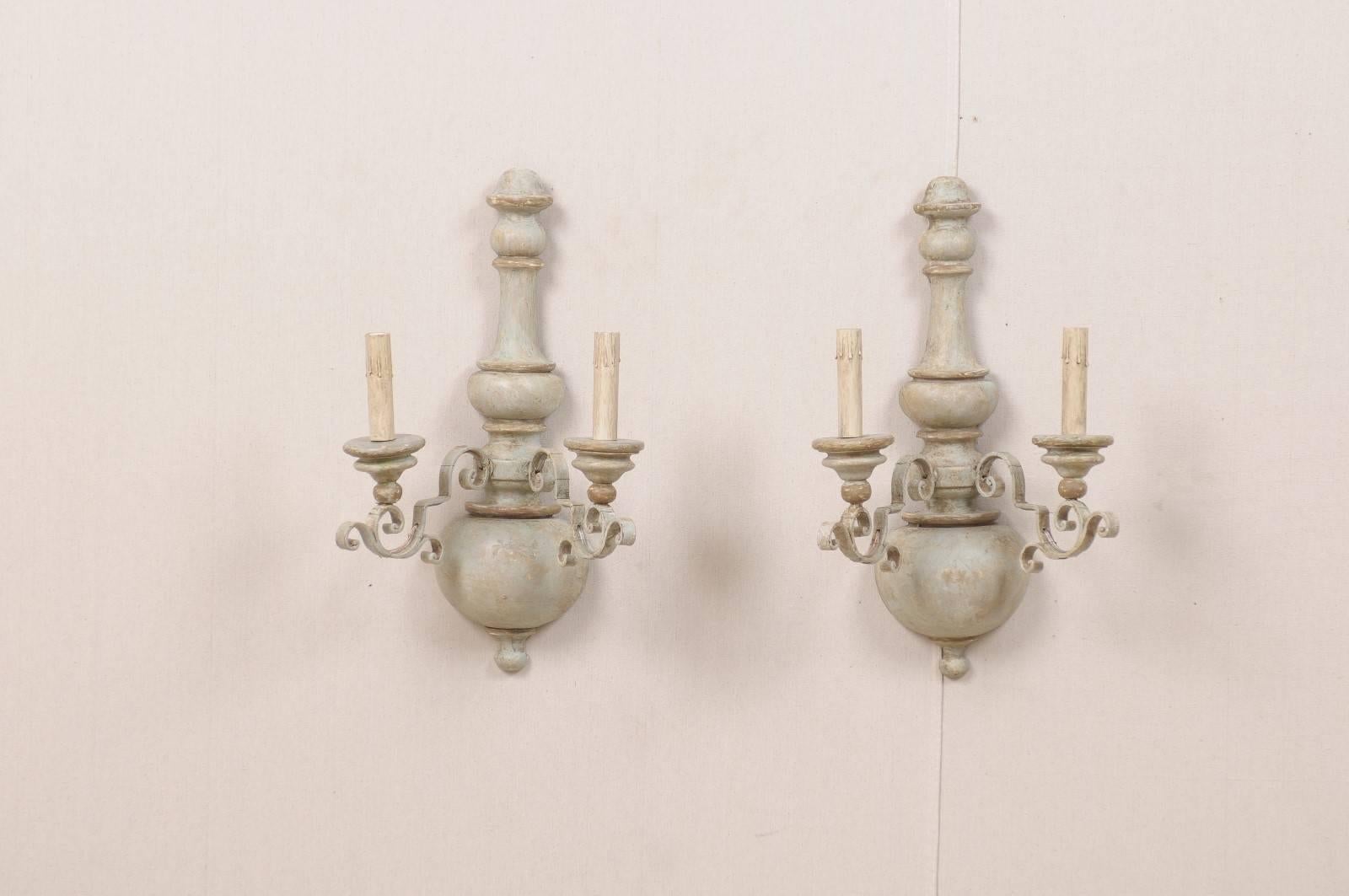 A pair of Italian painted metal and wood two light sconces. These Italian sconces are made up of turned wooden central columns from which two ornate metal scroll arms with lights emerge. The colors in this pair of sconces include light blue, green,