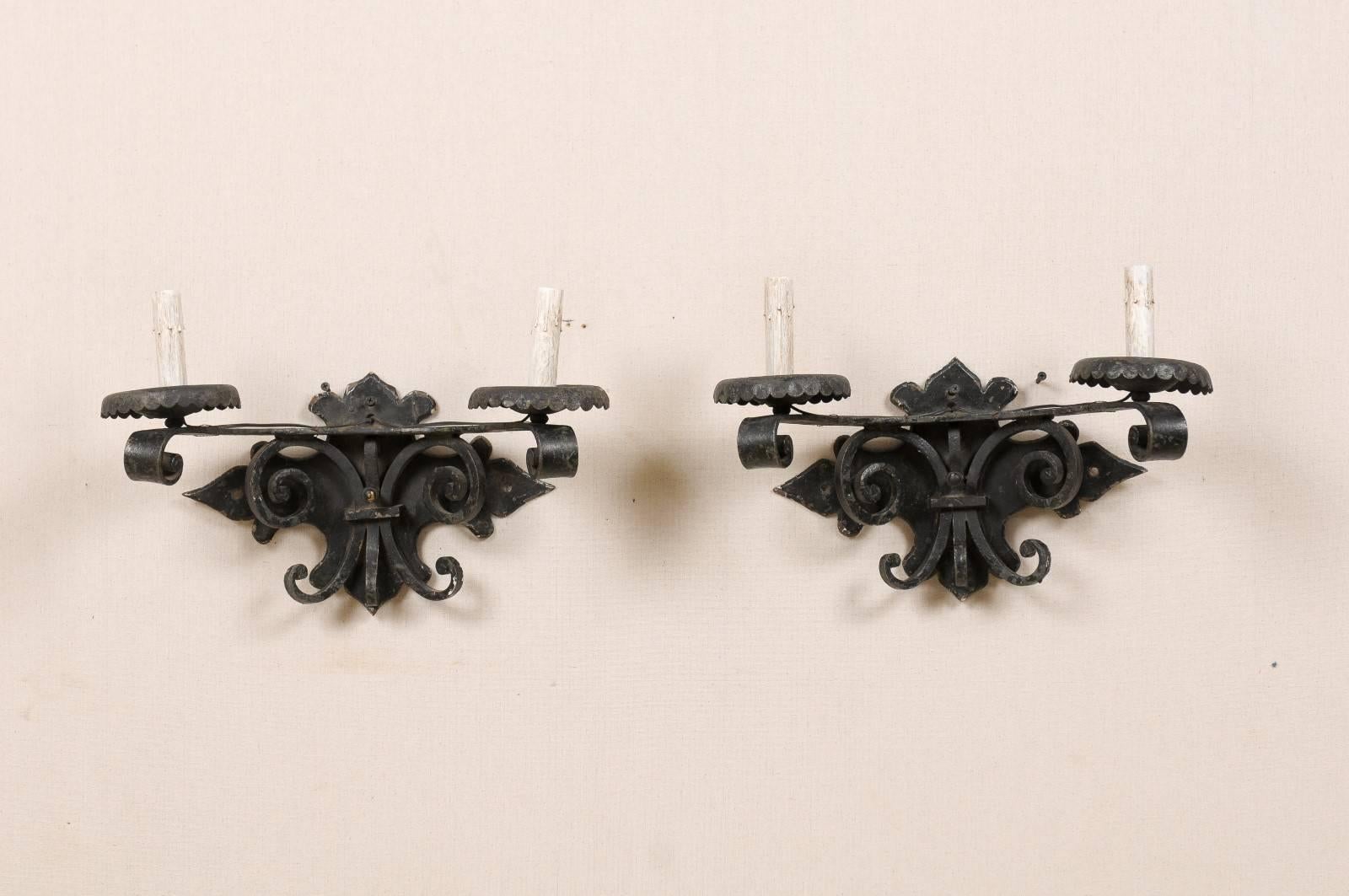 A pair of Spanish vintage dark colored forged iron two-light sconces from the mid-20th century. This pair of sconces is adorned with scrolls underneath the arms attached to a nicely shaped backplate. The scrolled arms support the metal bobèches and