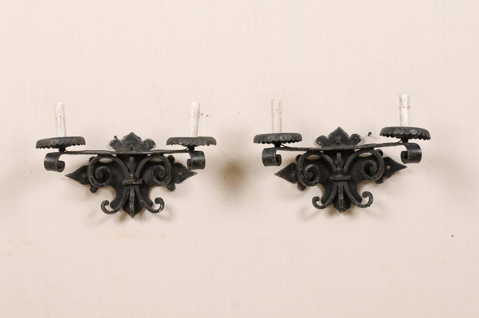 Paire d'appliques à deux lumières en fer forgé de couleur foncée d'époque espagnole, datant du milieu du 20e siècle. Ces appliques sont ornées de volutes sous les bras, attachées à une plaque arrière joliment façonnée. Les accoudoirs à volutes