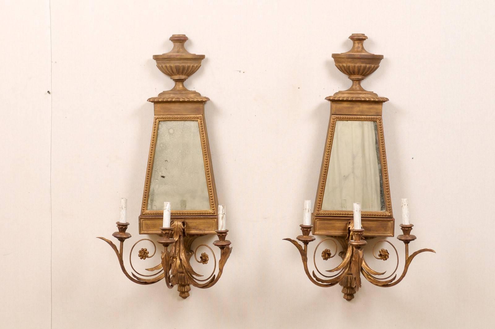 A pair of Italian vintage mirrored three-light sconces from the late 20th century. These Italian sconces are painted wood and metal and feature carved urn motifs at the top, scrolled arms with acanthus leaves motifs and mirrored back plates. The