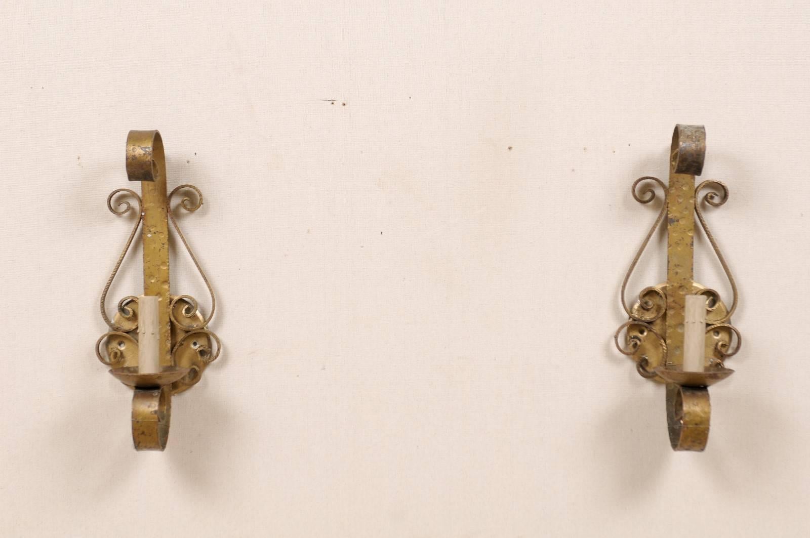 A pair of Spanish forged iron, painted gold single-light sconces from the mid-20th century. This pair of sconces has scrolled arms supporting the bobèches and painted candle sleeves as well as scroll decorations adorning the sides. This pair is a