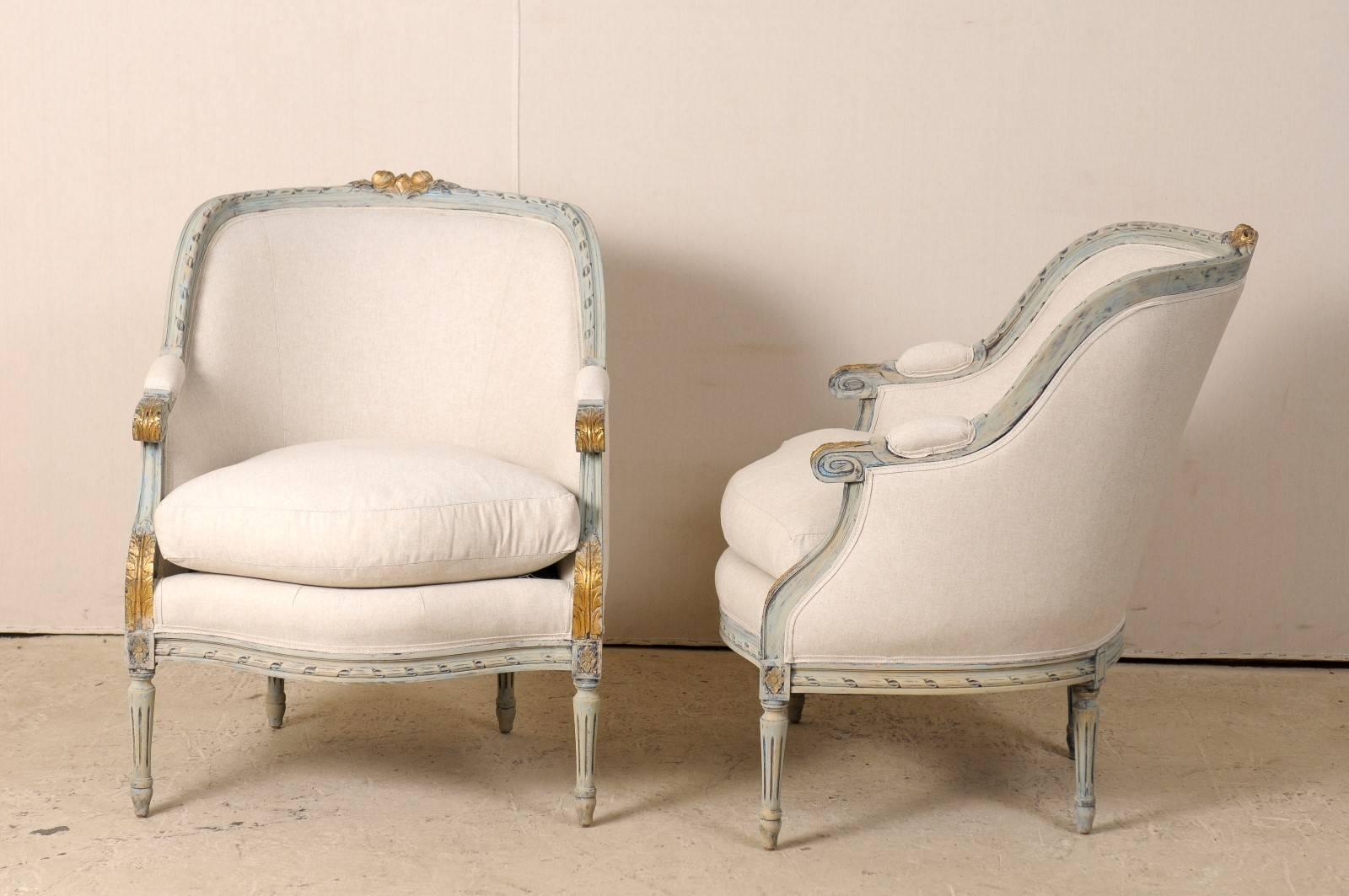 A pair of French Louis XVI style occasional barrelback bergères chairs, circa 1920. This pair of French painted wood chairs features scrolled and upholstered arms, fluted legs with rosettes on the knees, acanthus leaves and rose carvings on the top