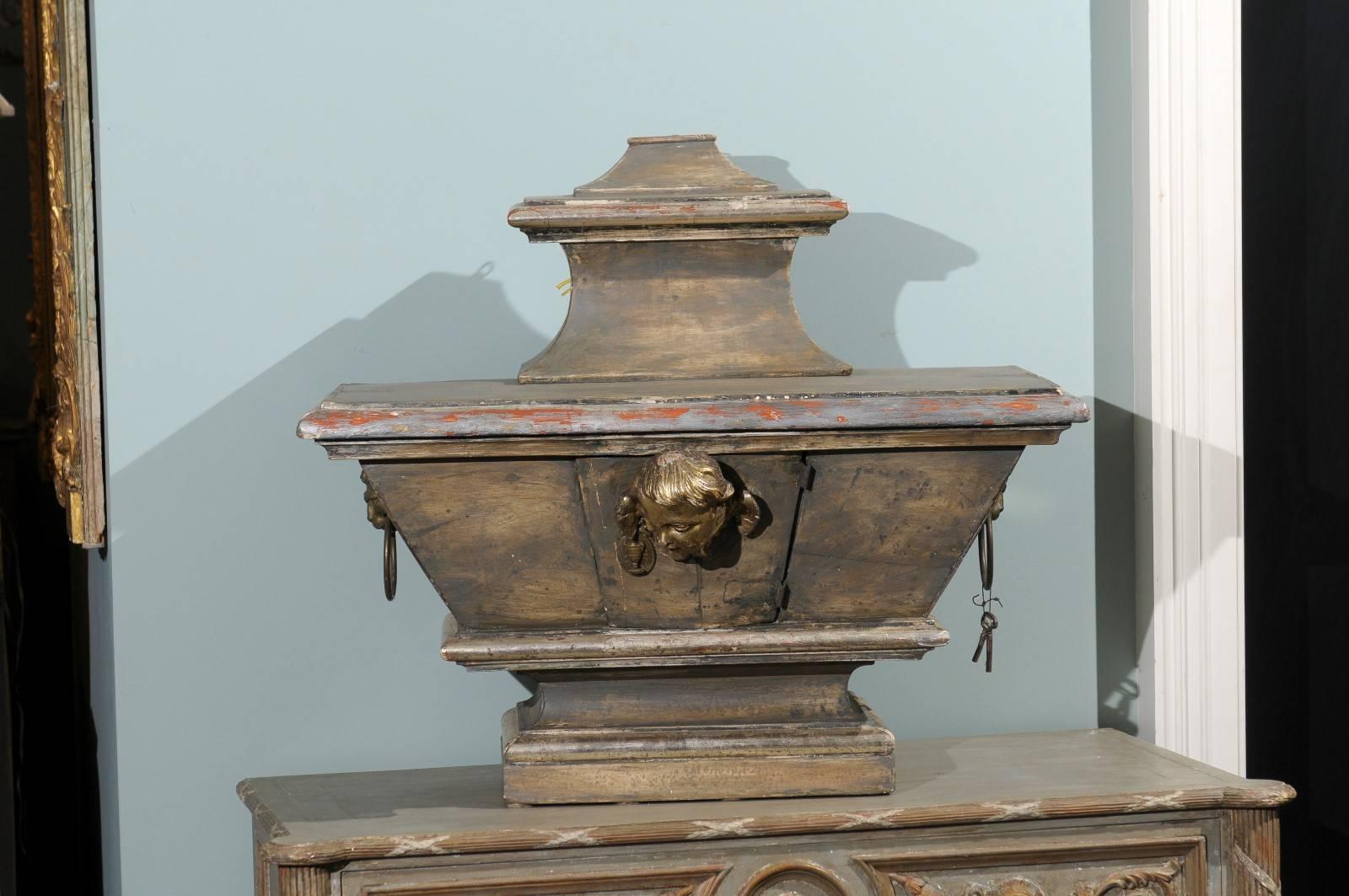 An Italian 19th century painted reliquary. This reliquary features a gilded and carved angel face on the door. The sides are flanked with and lions faces with ring pulls. The general color of the piece is more of a dark grey with very subtle warm