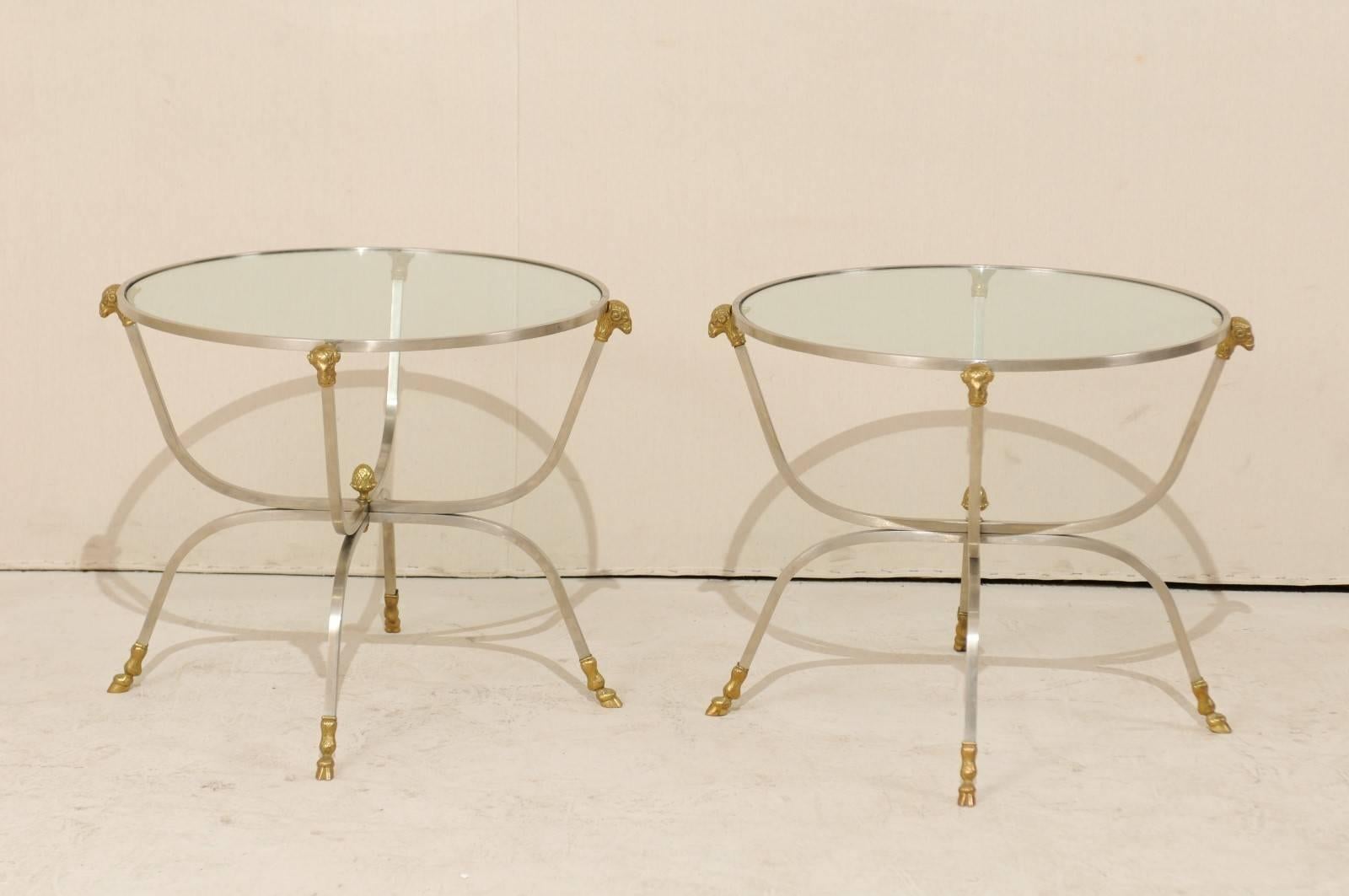 A pair of vintage Italian glass top occasional tables. This pair of Italian coffee tables from the mid-20th century feature round, glass tops, brass ram's head accents and hoofed animal feet. The frames are a silver metal and are made in a higher
