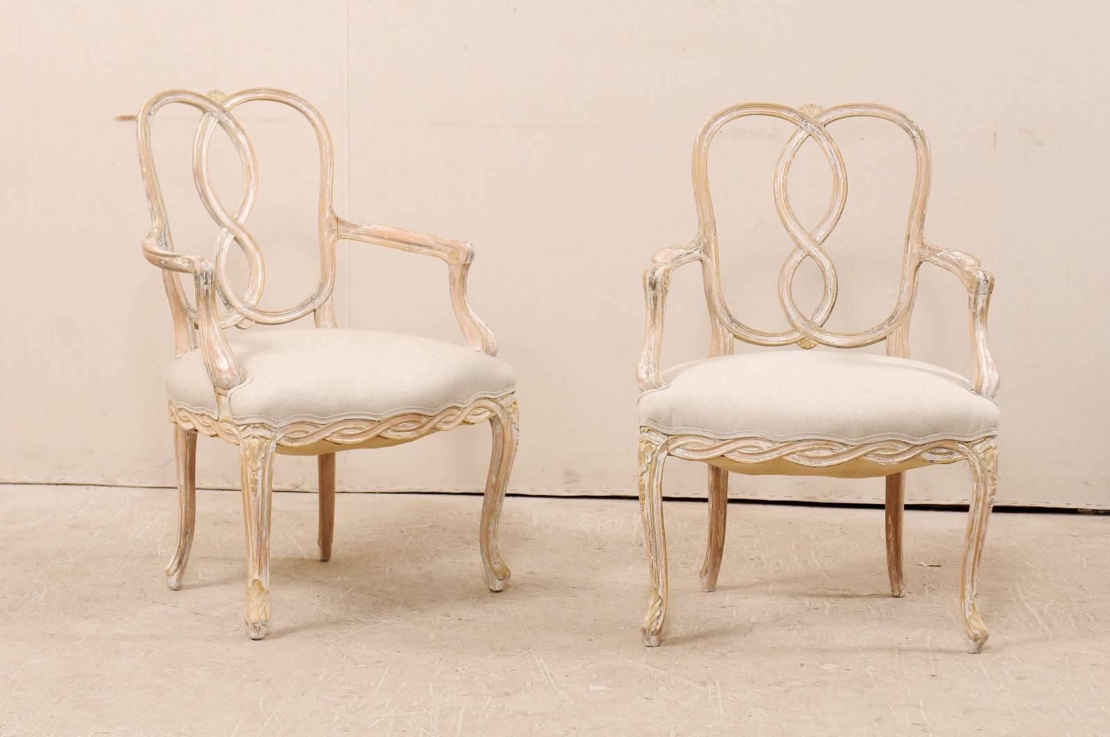 A pair of American made, Italian Venetian style painted wood chairs from the mid-20th century. This pair of armchairs feature a twisted ribbon motifs on their backs and skirts. The chairs have cabriole legs which are adorned in a leaf motif and the