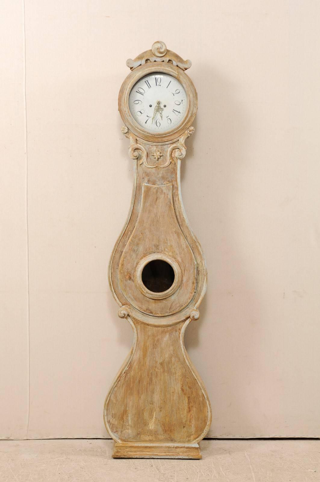 A 19th century Swedish Fryksdahl longcase clock. This Fryksdahl clock features a carved crest, volute accents at the neck with a central star, a round face, and a volute accentuated waist. This clock retains it's original metal face, hands and