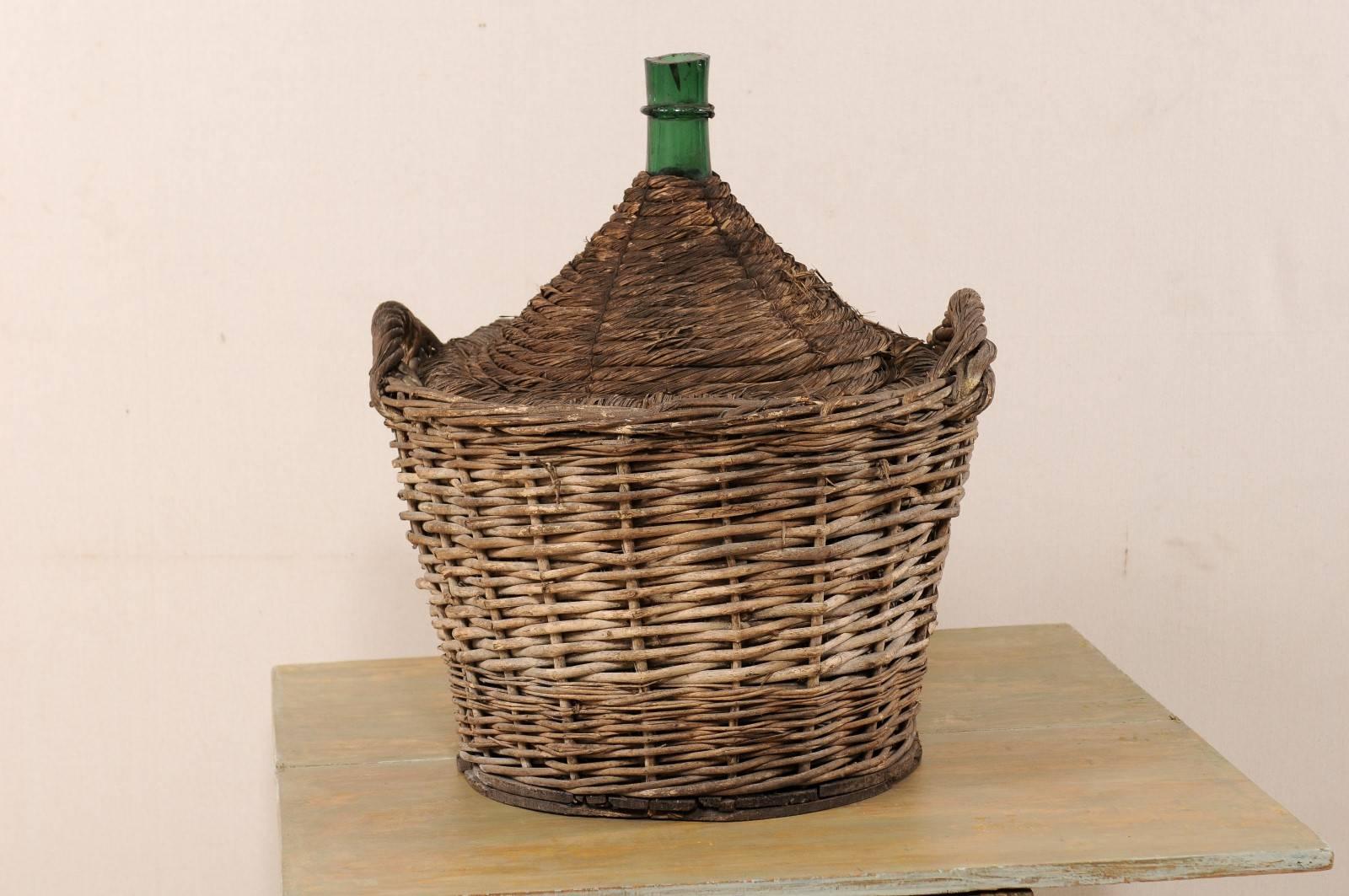 A Mid-Century French handwoven basket with demijohn wine bottle. This is a French two handle basket containing a large sized, green, demijohn wine bottle. The basket is hand woven and nicely aged. This French basket and demijohn bottle from the