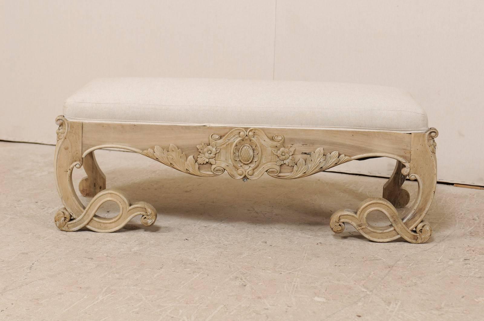 A Swedish carved wood and upholstered bench. This ornate Swedish wood bench from the early 20th century has an ornately carved skirt and legs with upholstered seat. The skirt features a scalloped front with central floral motif. The legs appear to