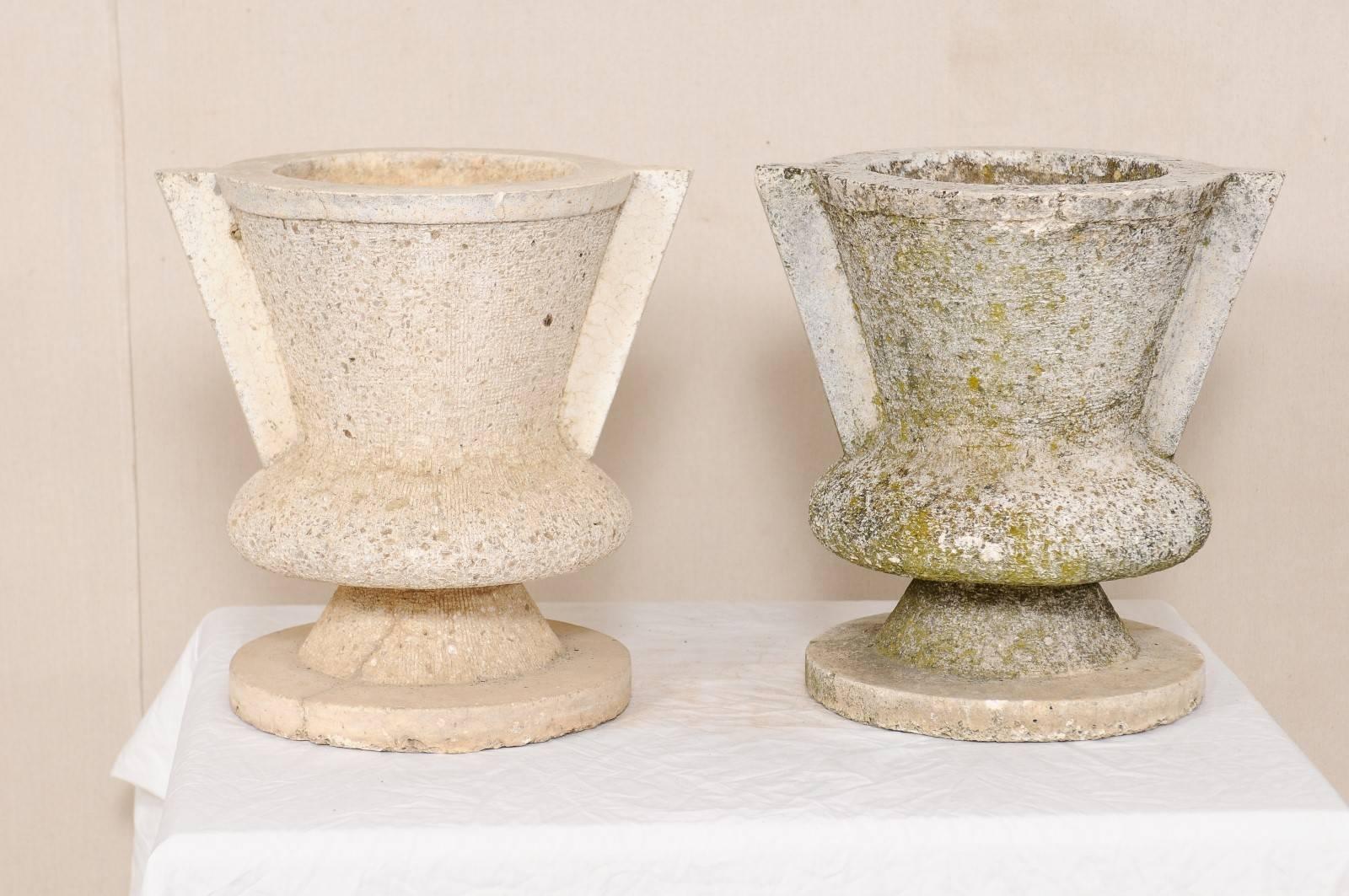 A pair of French urns. This pair of vintage French planters have been made of cast stone and have a modern design element to them. These shapely urns have a solid handle piece at either side, and are raised up on circular platform bases. These urns