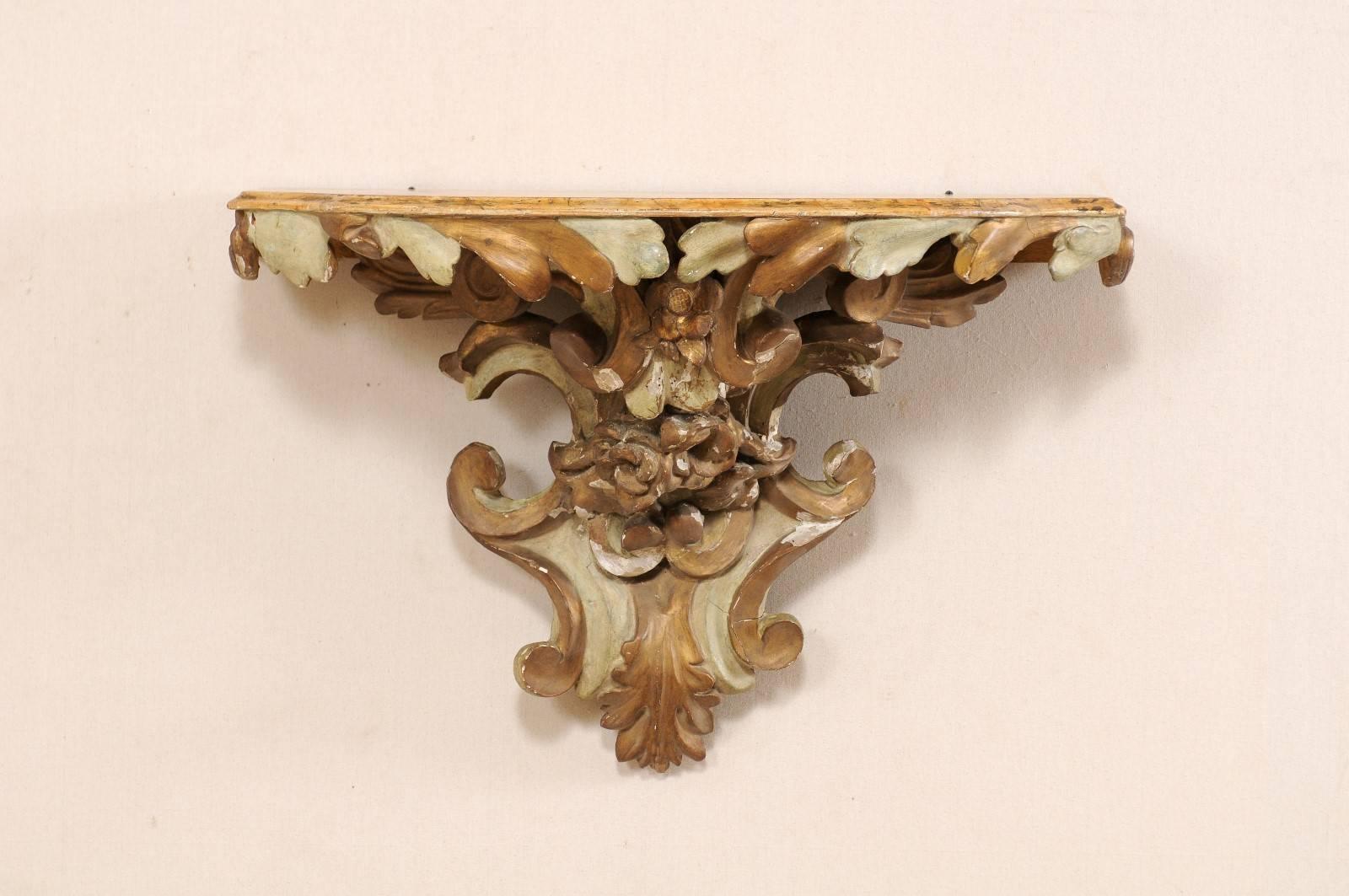 An antique Italian carved wood wall-mounted shelf. This Italian architectural piece, from the early 19th century, has been hand-carved in a scroll and leaf motif. The shelf top has a beveled edge and a faux marbled finish. The base is primarily a
