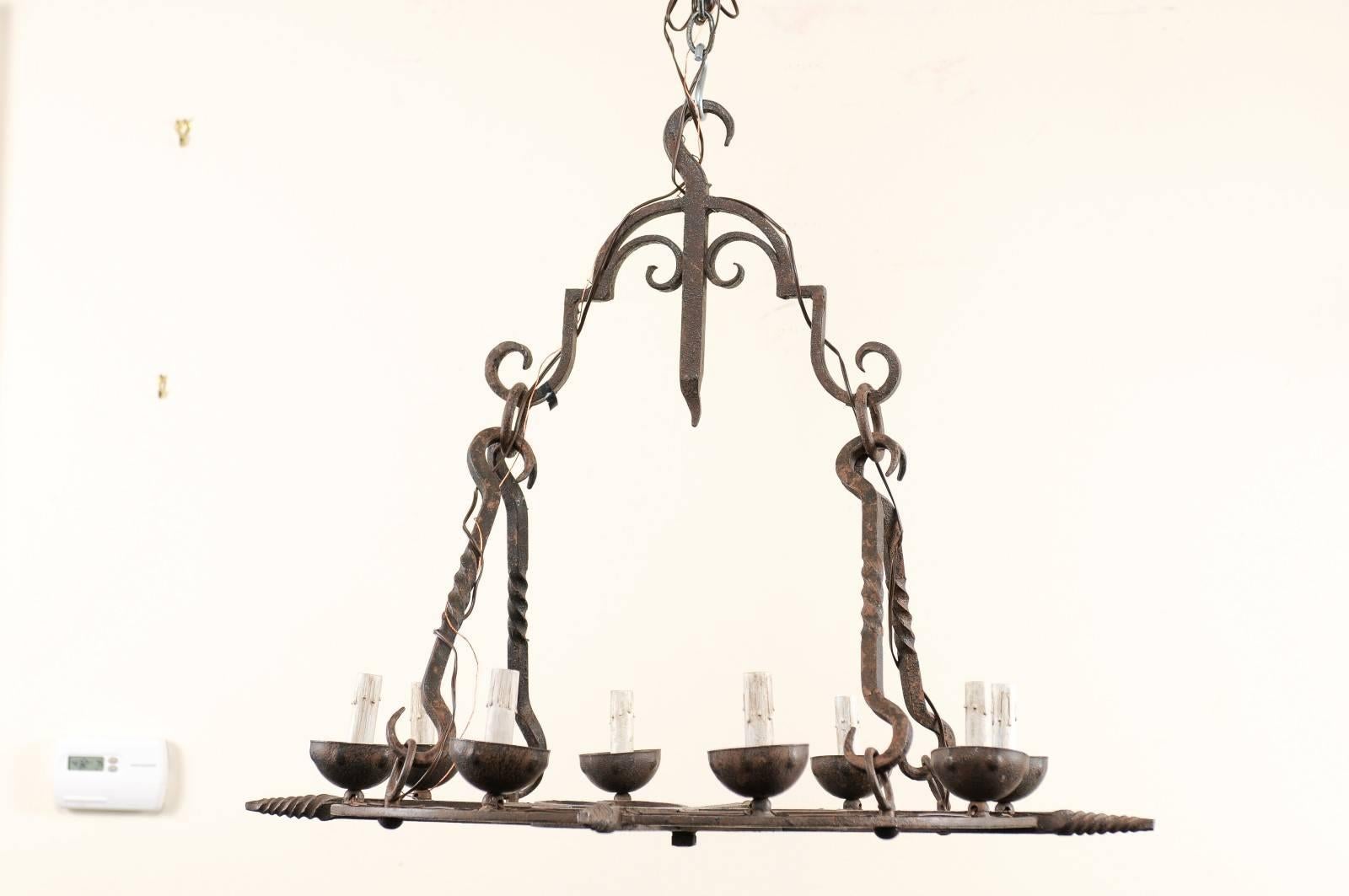 A French mid-century eight-light chandelier. This French chandelier from the mid-20th century of forged iron has a flattened, circular shaped body which supports the eight iron bobèche cups and candle covers around its perimeter. The lower section