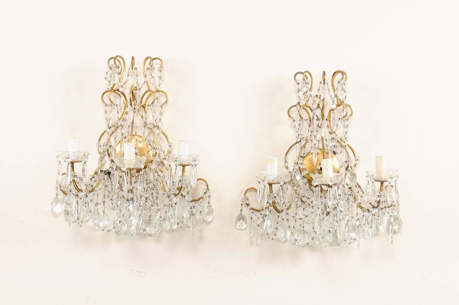 A pair of Italian vintage three-light crystal sconces. This pair of Italian crystal sconces each feature a variety of faceted crystals ornately decorating the scrolled and gilded metal armature and arms. Strands of crystals are heavily draped