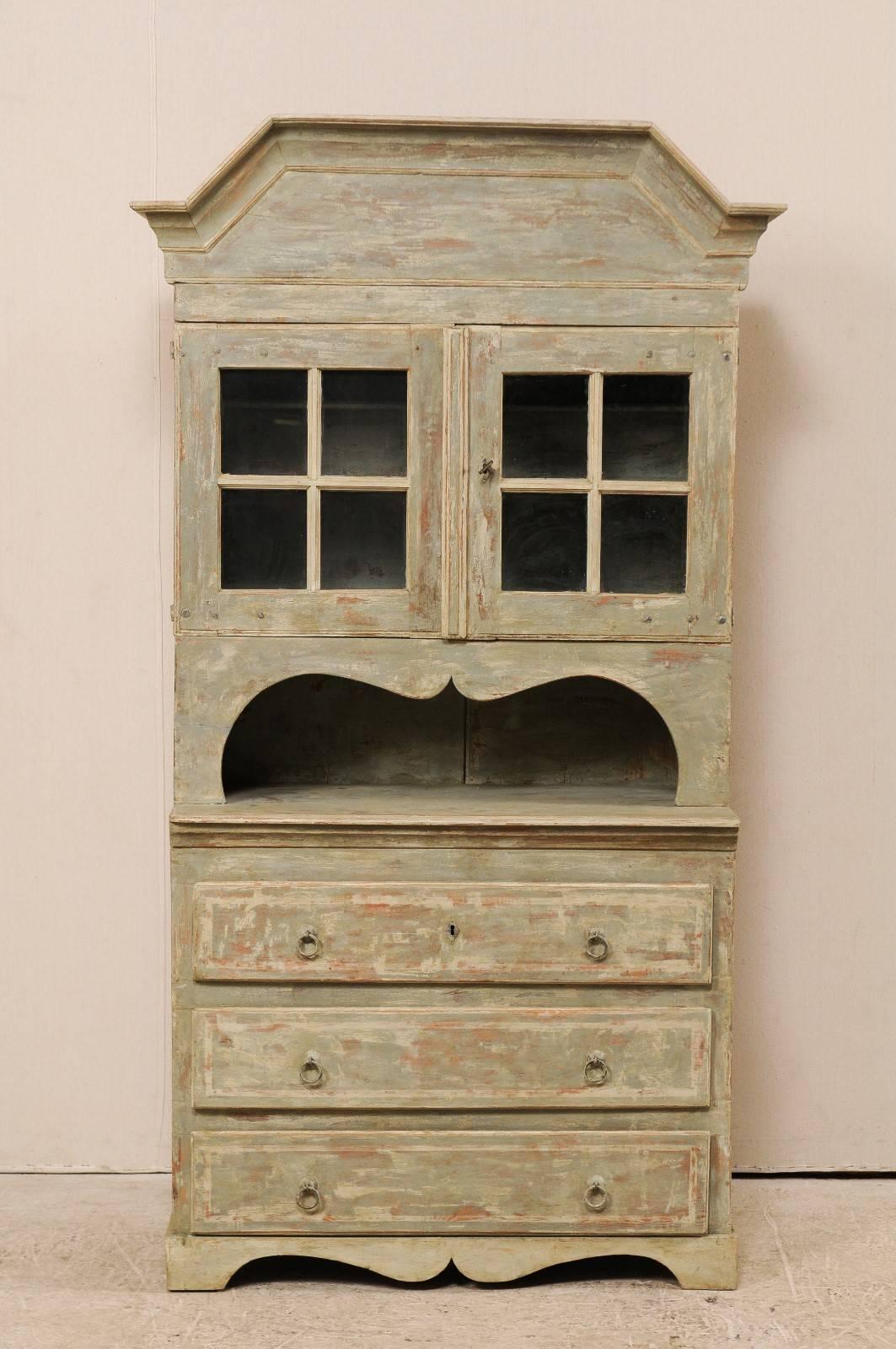 An early 19th century Swedish tall cabinet. This antique Swedish cupboard has a heavily molded pediment bonnet. There are two glass doors in the upper case, which sits recessed back over the lower case, comprised of three drawers. There is a shelf
