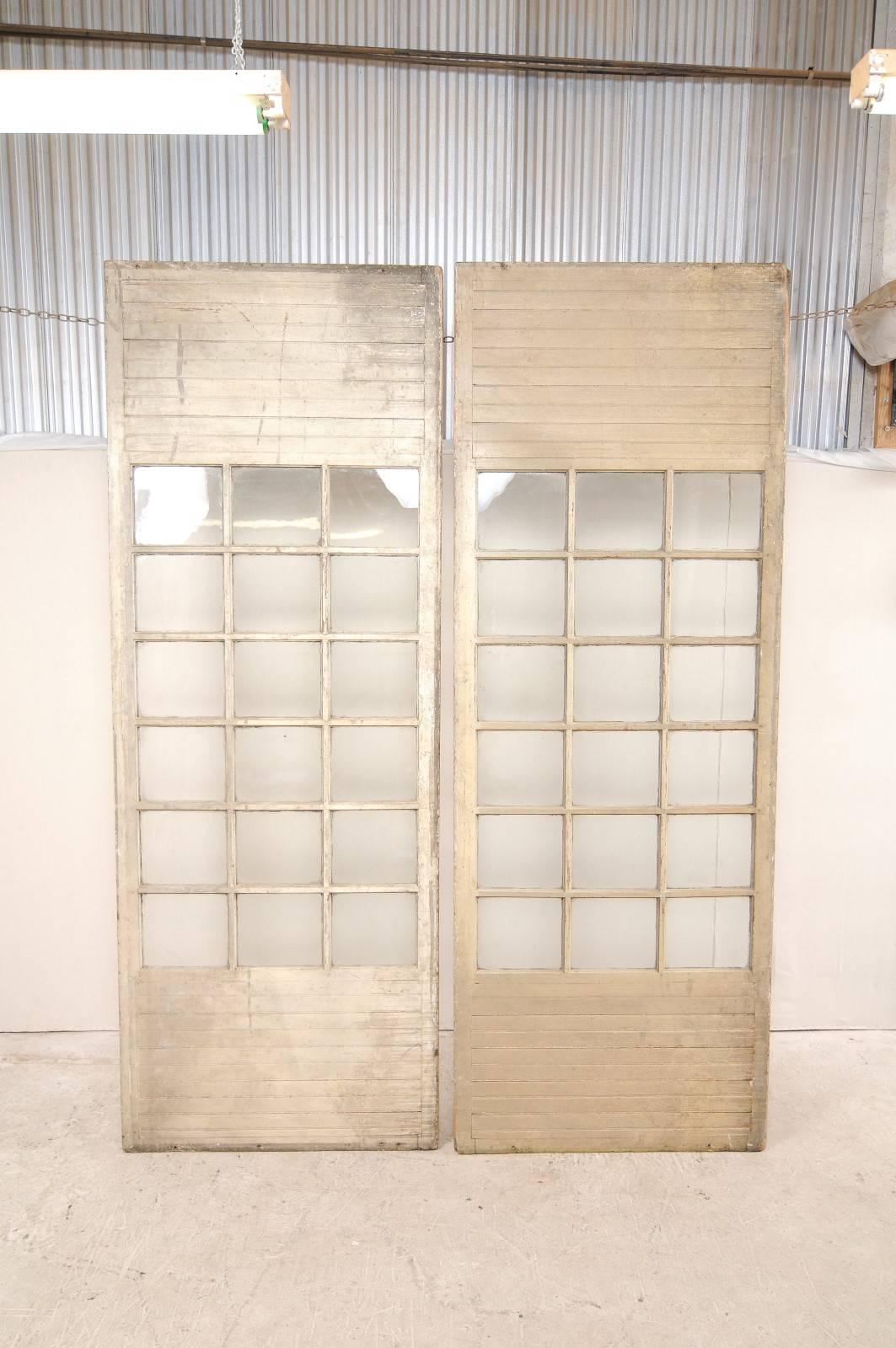 This is a pair of over-sized French doors from the early 20th century. These antique French doors are substantial, standing at a height of more than 10 feet! Each door has 18 glass panels, framed by vertically slatted wood at the upper and lower