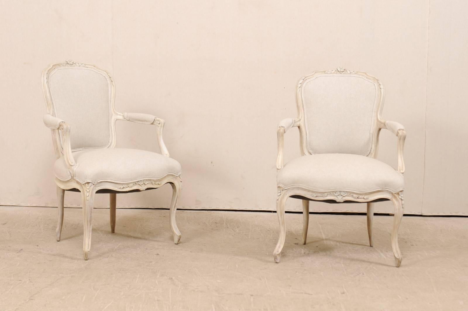 A pair of Swedish Louis XVI style armchairs from the mid-20th century. This pair of Swedish upholstered and carved wood arm chairs feature nicely carved floral motifs about the upper crest rail, center skirt and knees. The arms are padded at their