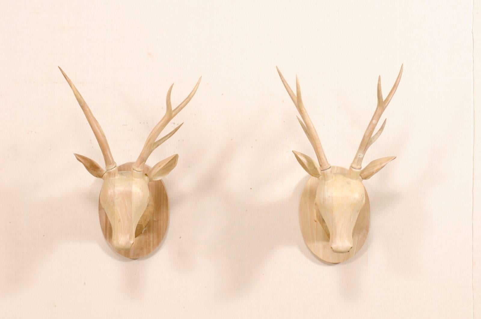 A pair of wall-mounted carved wooden deer heads. This pair of deer heads have been hand-carved of a bleached, tropical hard-wood in a custom run of two. The deer each have antler, are straight facing, and mounted onto oval carved wood back plates.