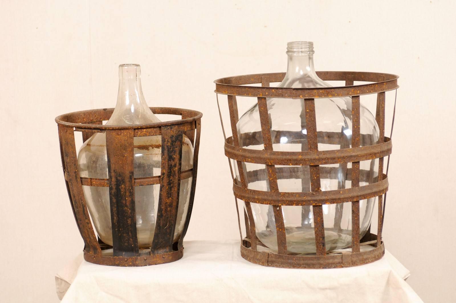A pair of midcentury French vintner iron baskets with demijohn wine bottles. This is a pair of French vintner iron-woven baskets, each containing a clear, demijohn wine bottle. The baskets are nicely aged with lovely patina. These French vintner