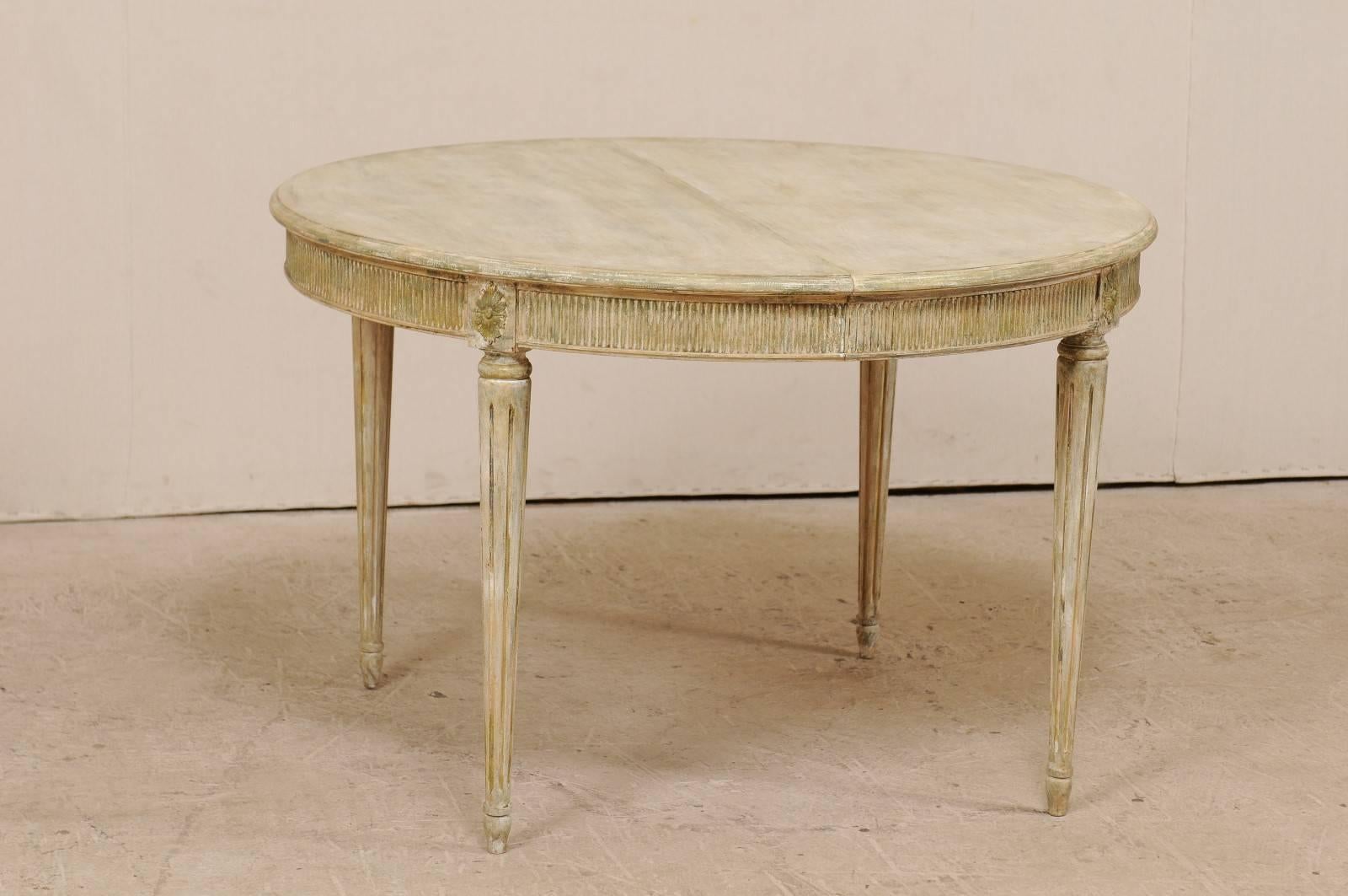 A vintage Swedish Gustavian style round painted wood table with two leaves. This Swedish table features a carved reeded apron and is raised on round, fluted and gently tapered legs which are accented with rosettes carved at the knees. This table