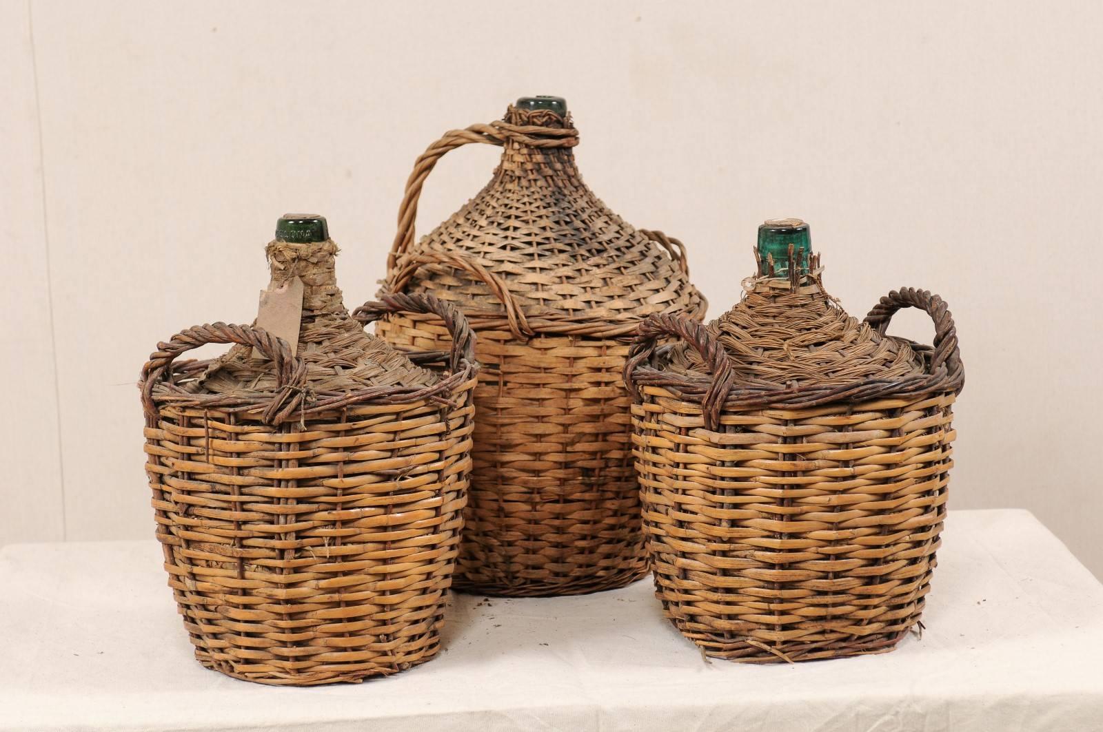 A set of three of midcentury French vintner woven baskets with demijohn wine bottles. This set of French vintner wicker-woven baskets, each containing a green colored, demijohn wine bottle. One basket is a two-handled, while the other two each have