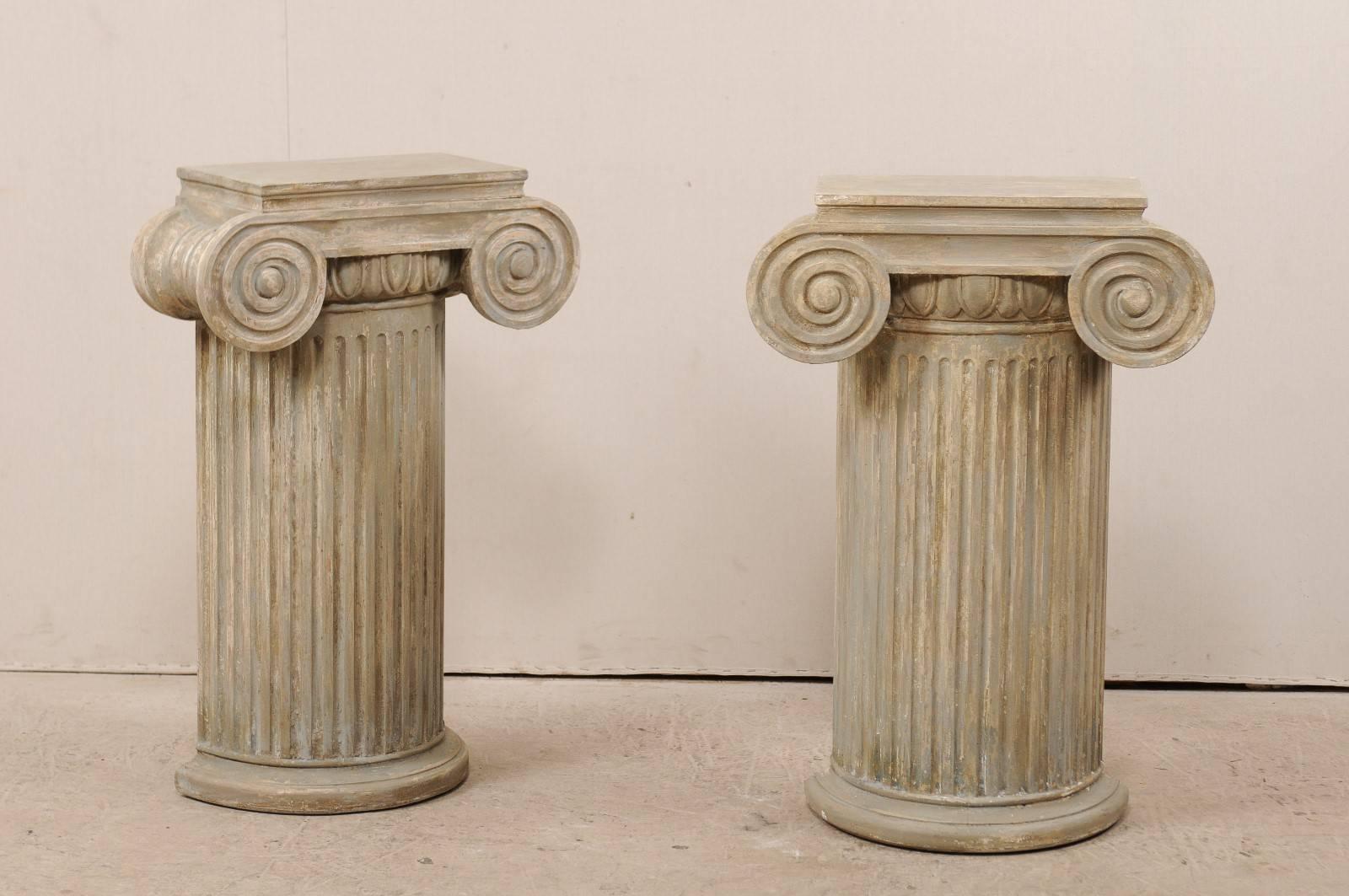 A pair of vintage American carved wood ionic column pedestals. This pair of pedestals are ionic style with rectangular-shaped tops over semi-rounded and fluted bodies, raised up on rounded bases. The overall color palette consists of various shades