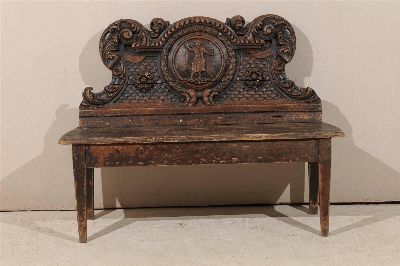 An Italian 18th century pinewood bench with richly carved ornate details. This dark wood Italian bench features a central medallion adorned with a man's figure. Around the central character and medallion are other beautifully carved details such as