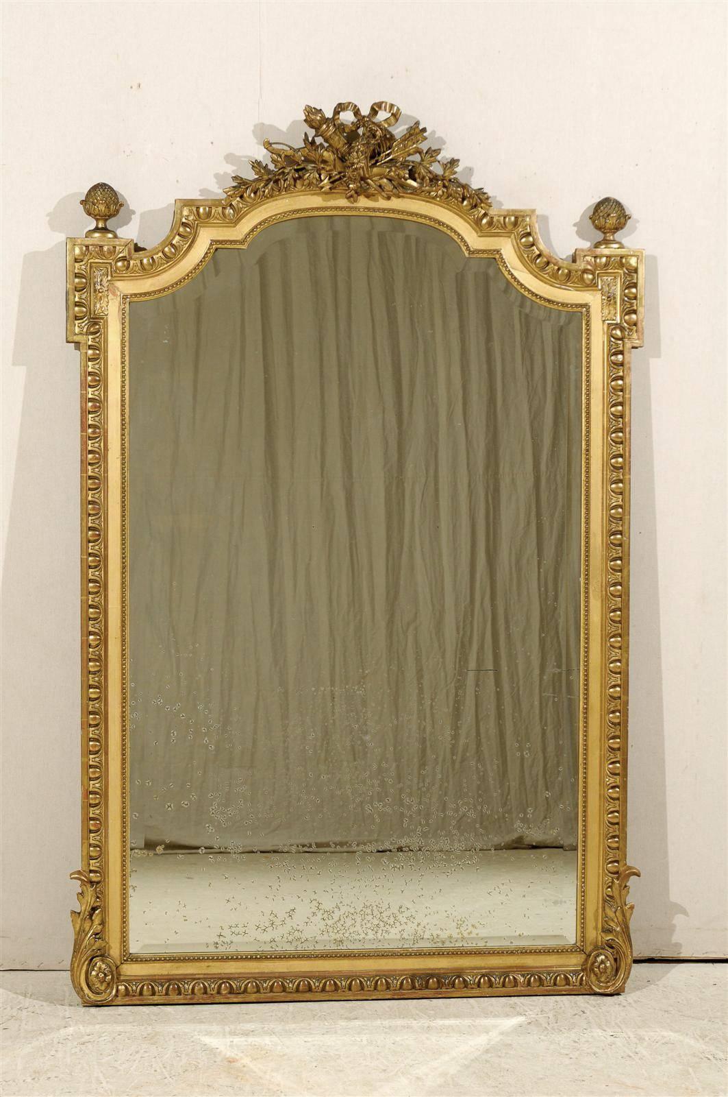 A tall French richly carved and gilt mirror from the early 20th century.  This large-sized gilded wood mirror features a crest elaborately adorn with a bow-tie, ribbons, arrows and torches, flanked within pine cone finials at each shoulder.  An egg