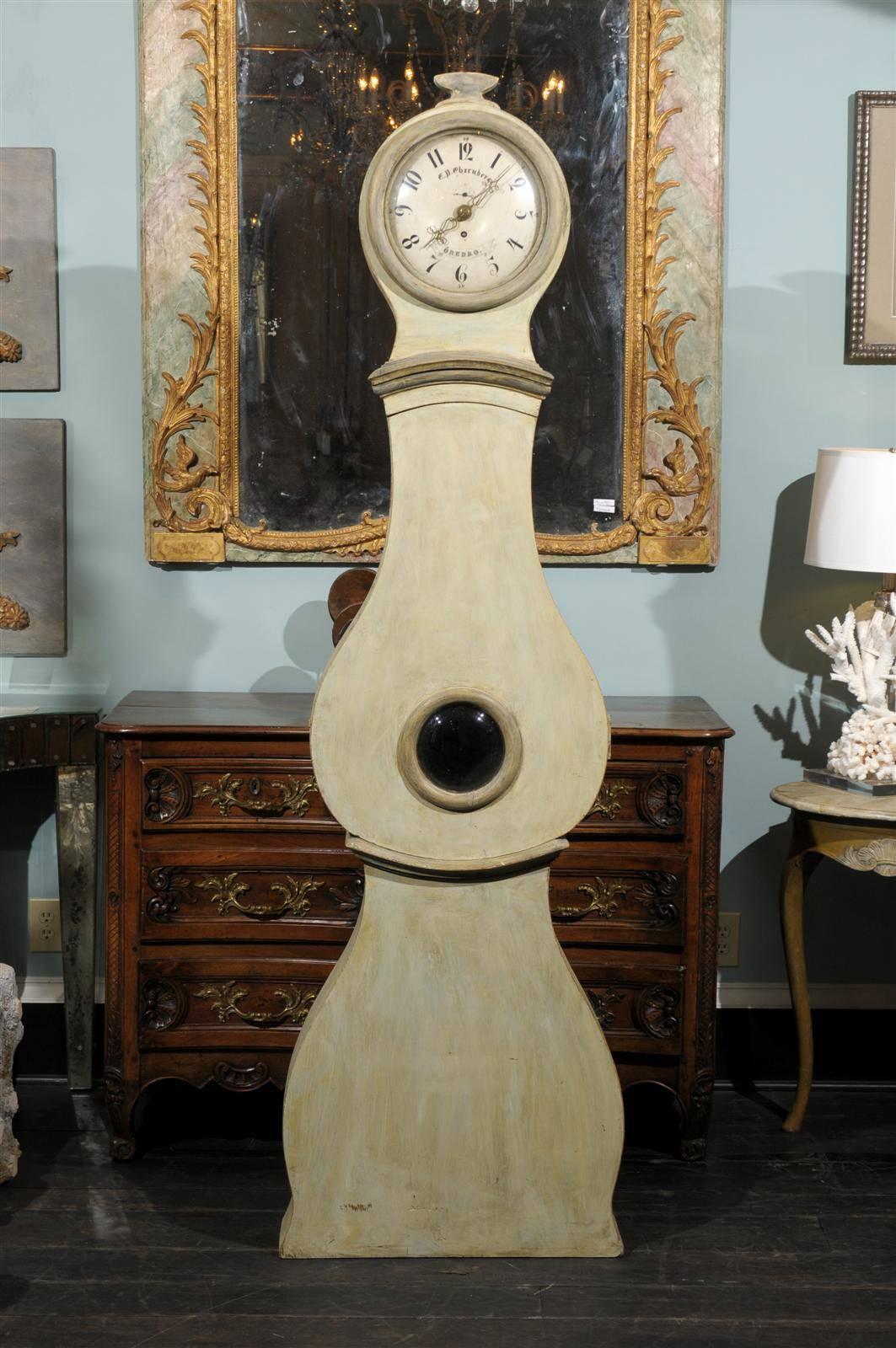 A 19th century Swedish painted wood clock with small carving at the crest, large round belly and flat base. Light grey/green color. This clock retains it's original metal face, hands and movement.  Signed E.P Ehrenberg. Orebro.

This clock would be