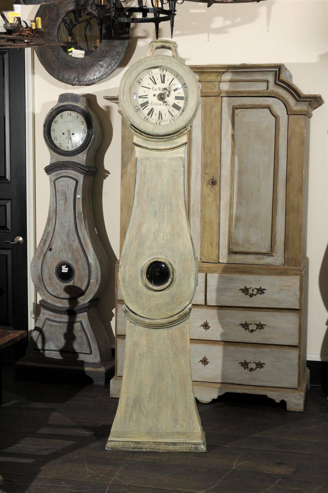 A Swedish 19th century painted wood clock. This clock retains it's original metal face, hands and movement.  This Swedish clock features a light grey or green color with hints of blue. The trims have dark grey tone. The clock is topped with a carved