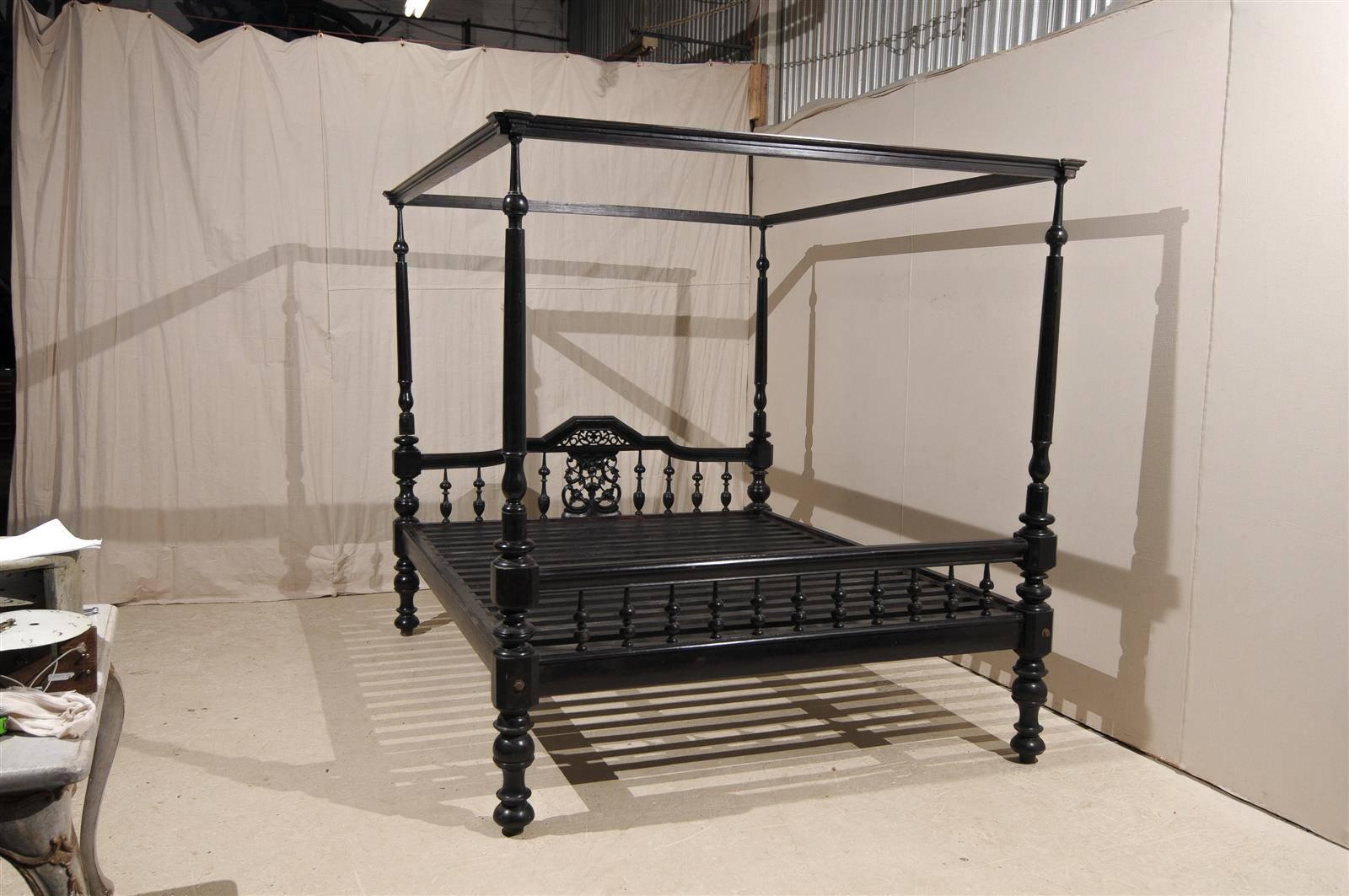 An antique anglo-indian black four post canopy style bed with turned legs and balusters. The headboard features some intricate carving with C-scrolls and foliage depictions in its center. The bed is also has some red undertone coming through.This