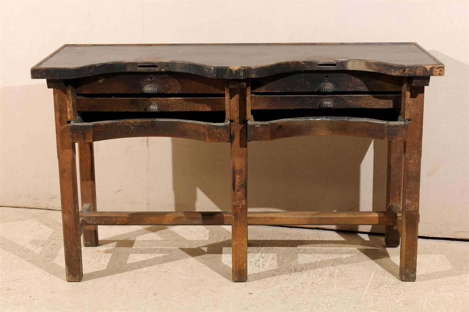 A Dutch wooden jeweler's work table. This jeweler's table is made of several drawers with flat fronts for the upper ones and concave ones in the lower section, echoing the top of the table. It is raised on straight legs joined by a cross and side