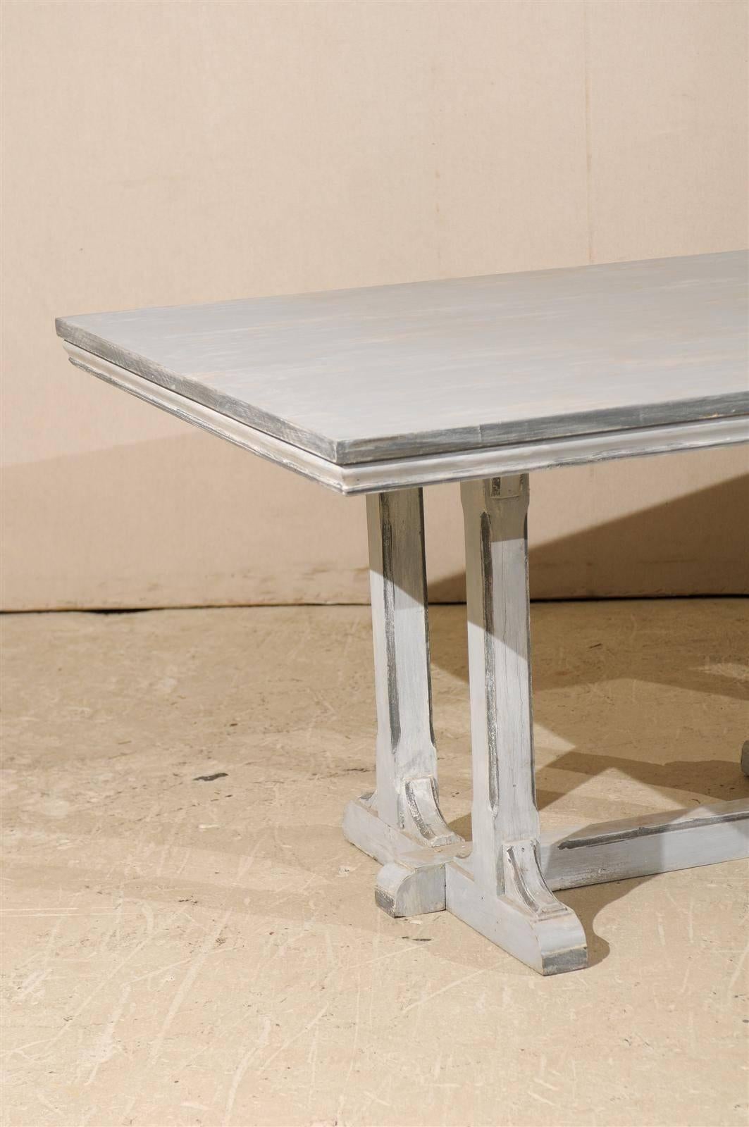 A large European grey painted wood rectangular dining table. This dining table features an old trestle base with cross stretcher supporting a later top. The edges have been highlighted with darker grey accents, providing more visual depth, 20th